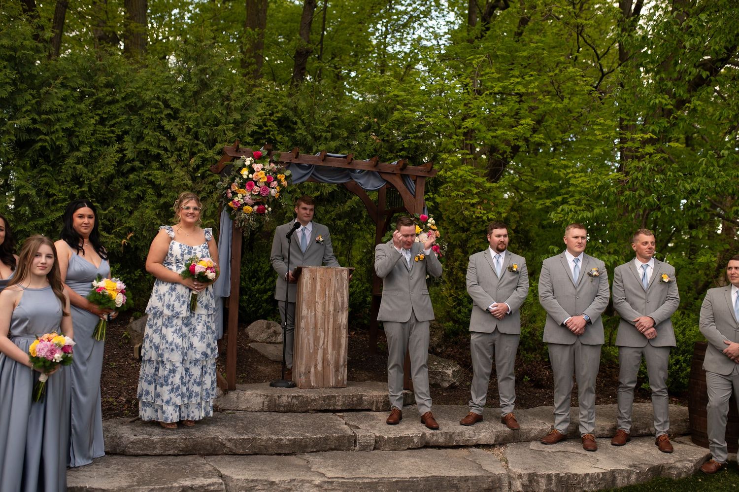 Wedding party in gray attire poses outdoors in a panoramic photo with lush green trees and natural scenery.