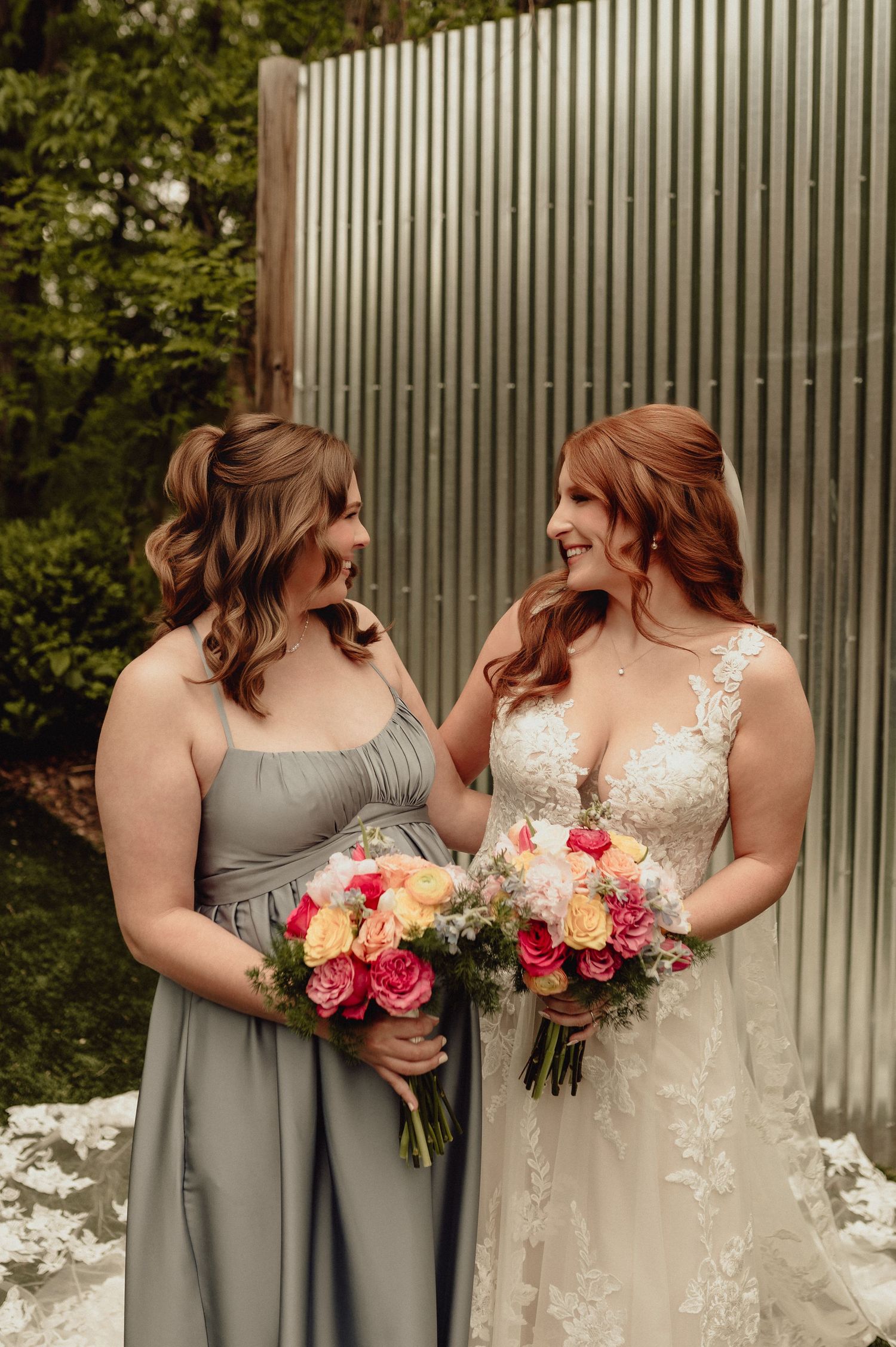 A bride in a white lace gown stands with bridesmaids in gray dresses holding vibrant pink and yellow floral bouquets.