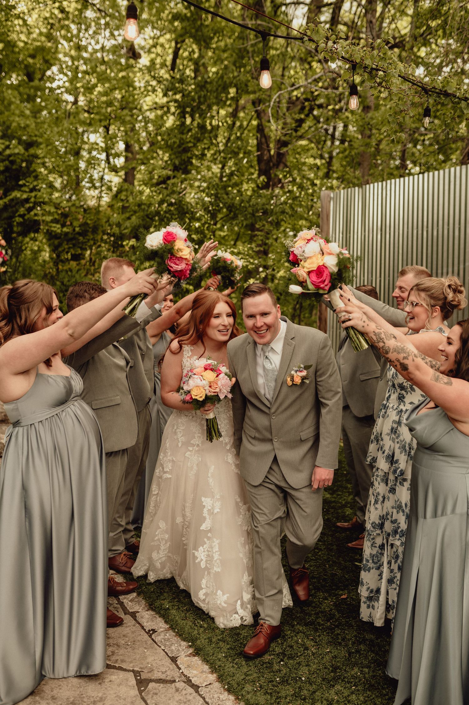 Wedding party in sage green dresses forms tunnel on garden path as couple walks through with floral bouquet.