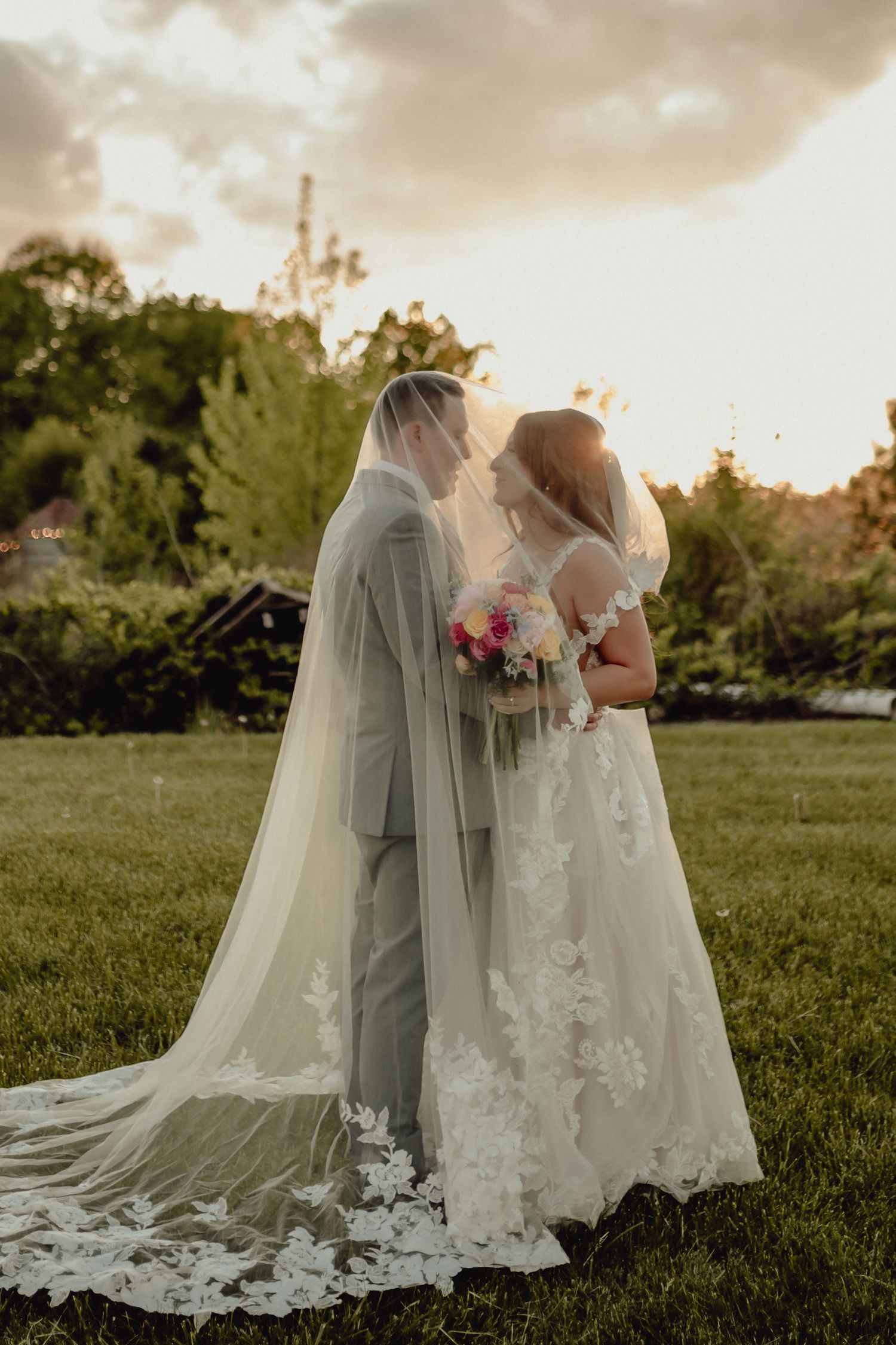 A romantic outdoor wedding photo at sunset with a couple in elegant white attire and flowing veils standing on grass.
