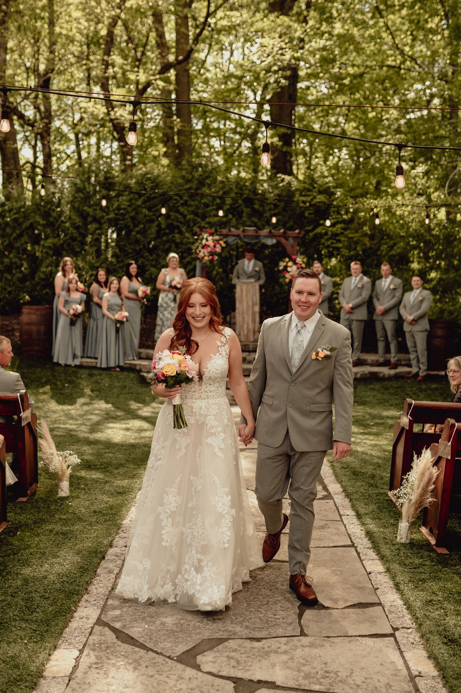A wedding ceremony takes place on a stone path surrounded by lush greenery and trees in a garden setting.
