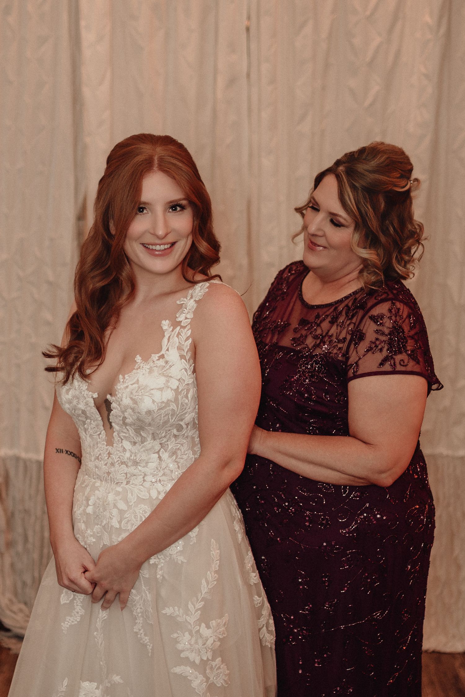 A bride in a white lace dress embraces someone wearing a burgundy sequined gown during a wedding celebration.