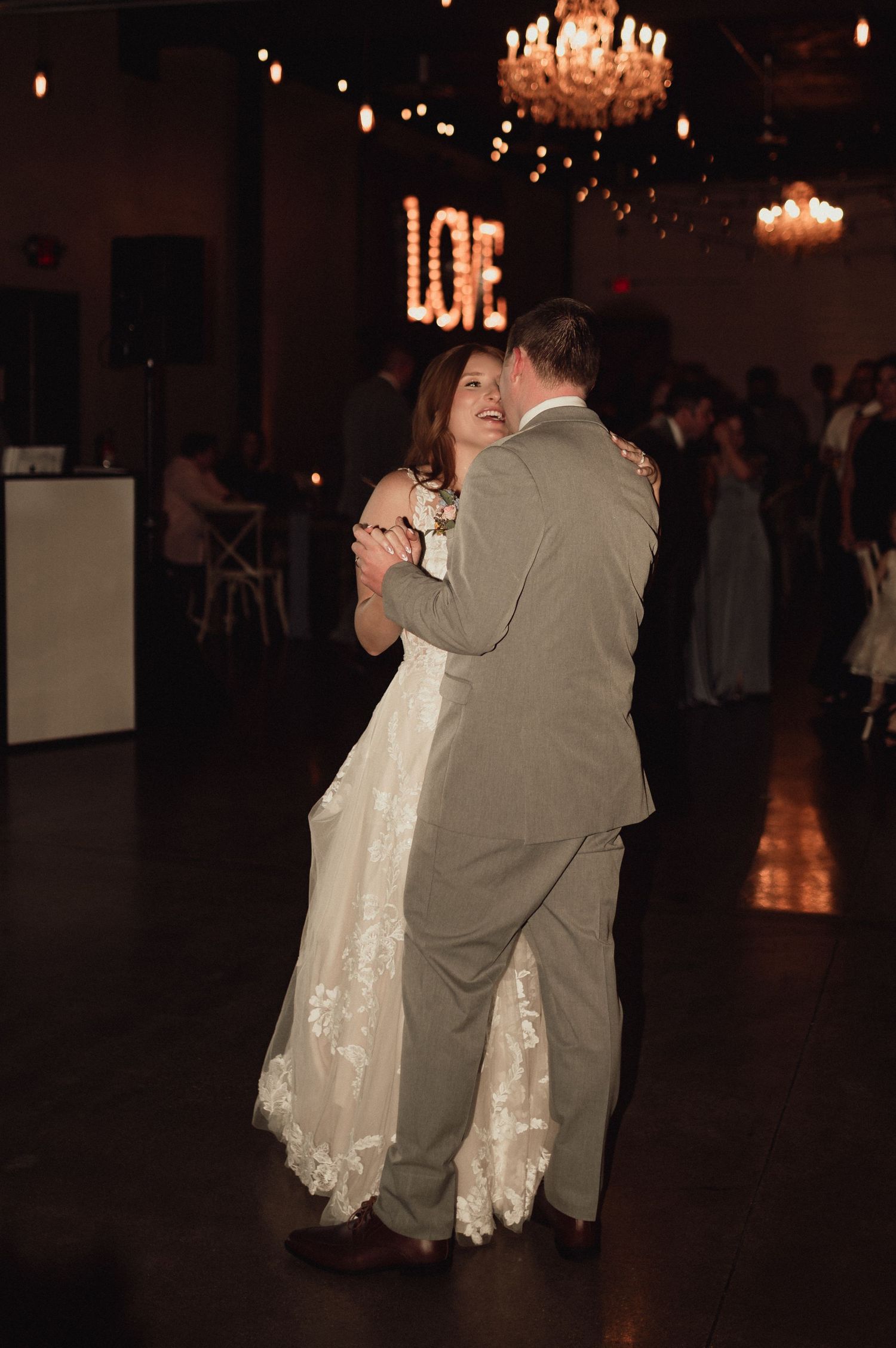A couple shares their first dance at a wedding reception under elegant chandeliers with a glowing LOVE sign in the background.