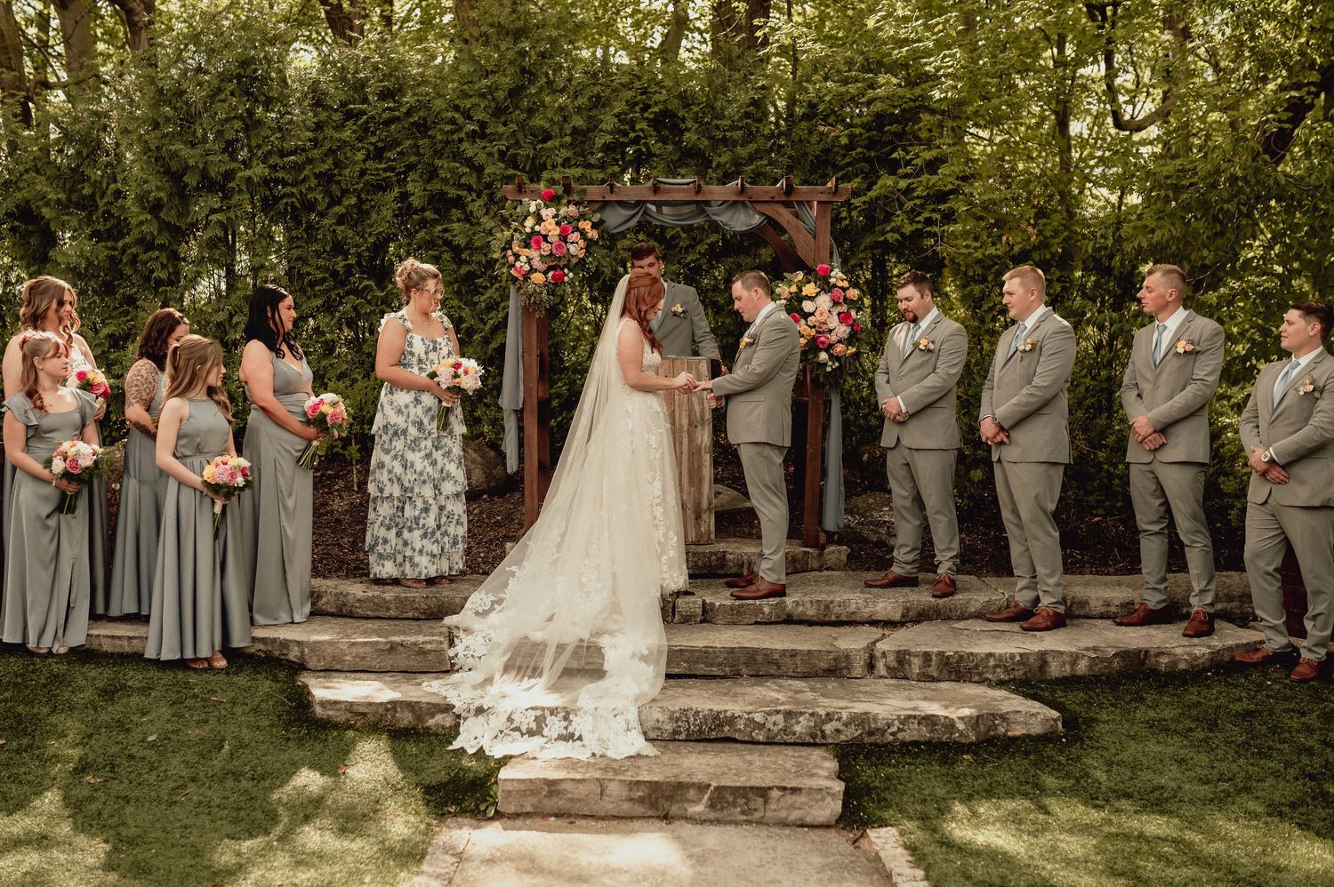 Wedding party in formal attire poses outdoors in a garden setting with stone steps and greenery.