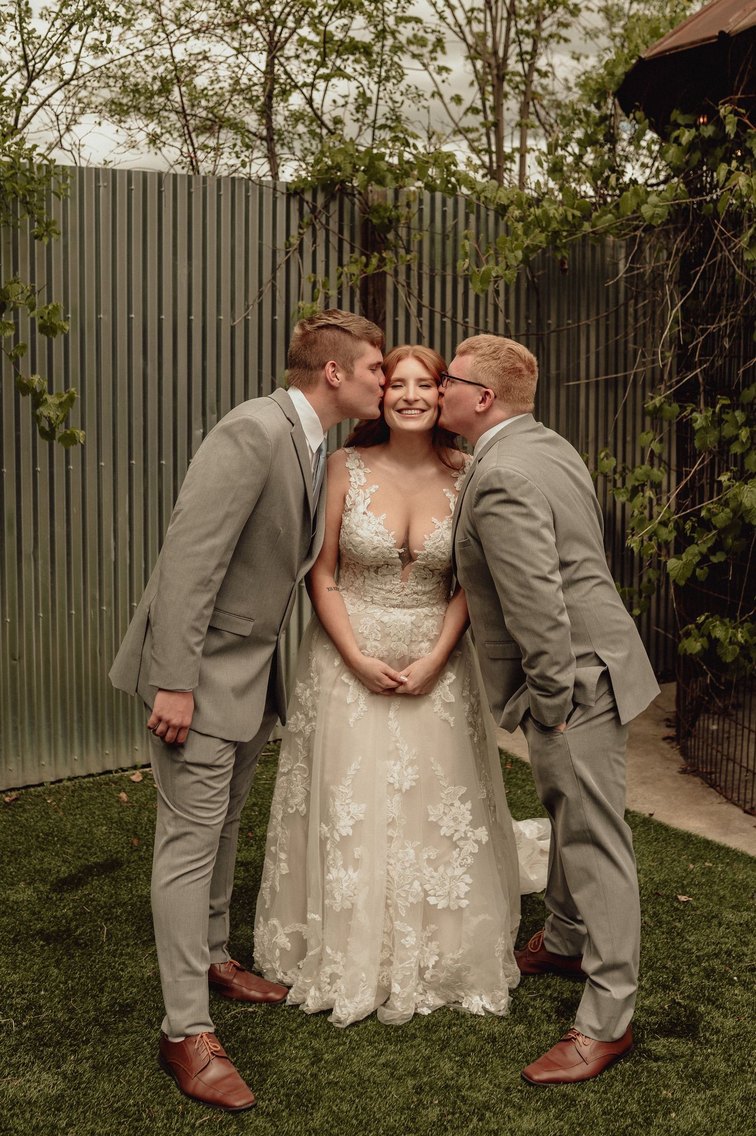 A tender moment captured outdoors with three people in formal attire on a grassy lawn with a fence backdrop.