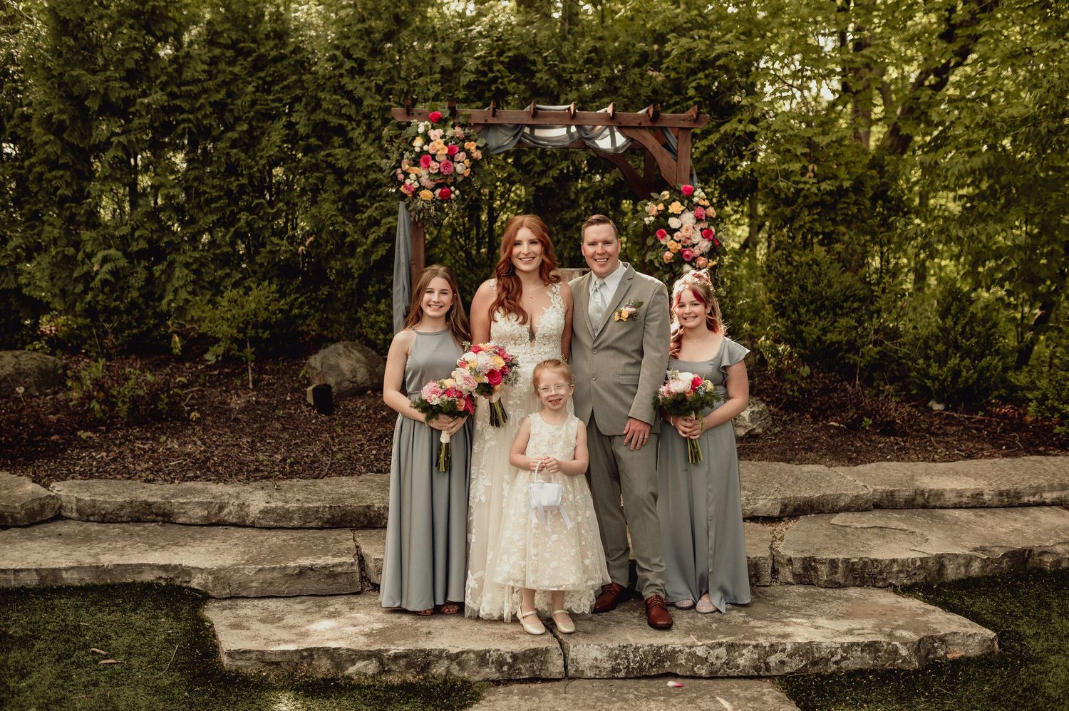 Wedding party in gray and white attire poses on stone wall in outdoor garden setting.
