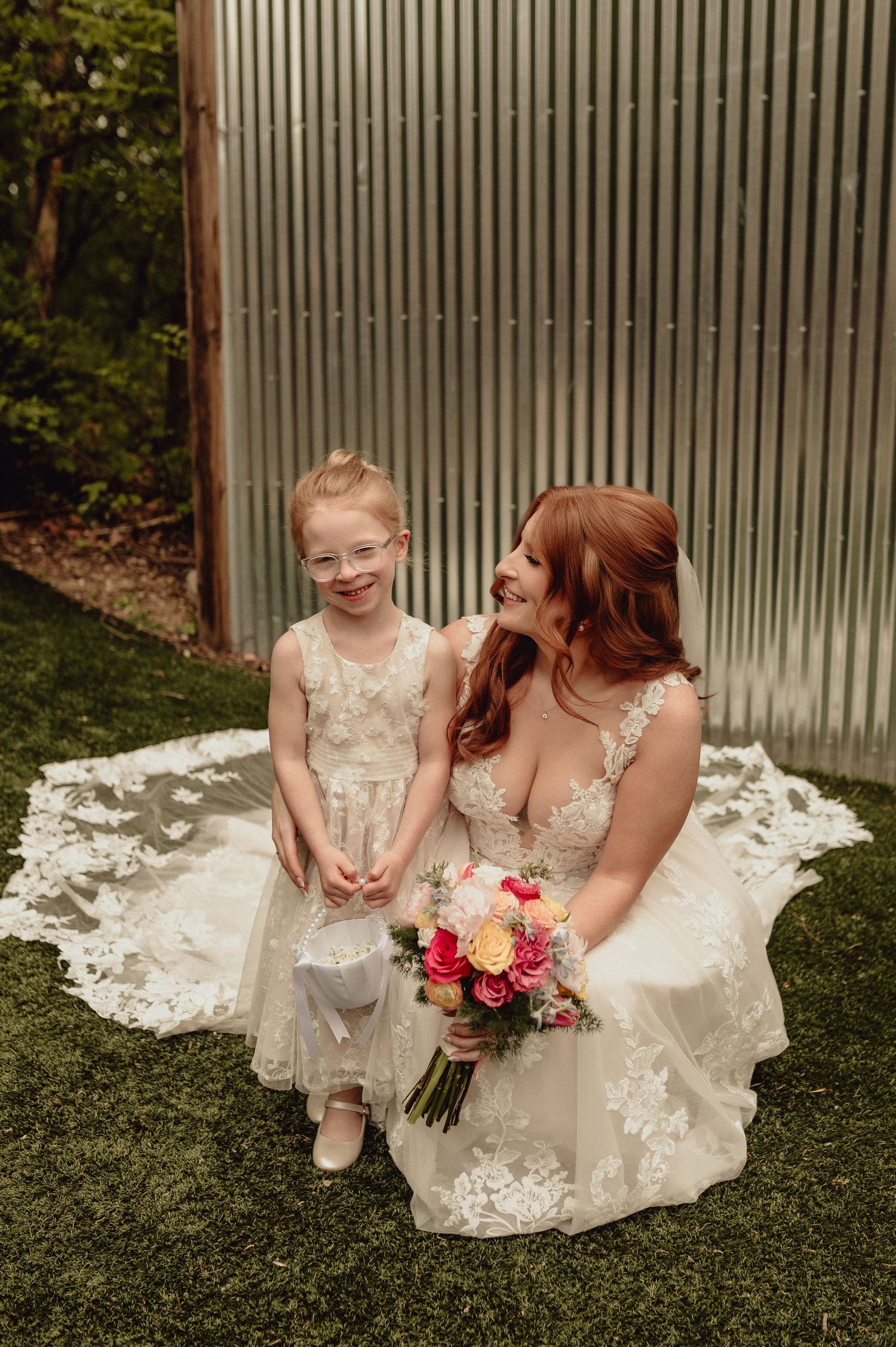 A tender moment shared between two people in white lace dresses sitting together on grass with a colorful bouquet.