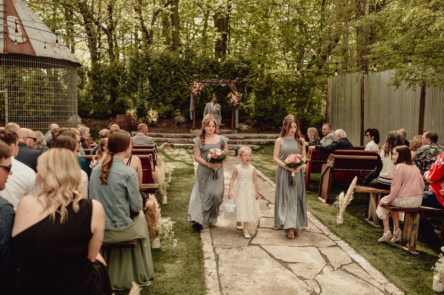 Outdoor wedding guests mingle and socialize at a rustic venue surrounded by greenery and trees.