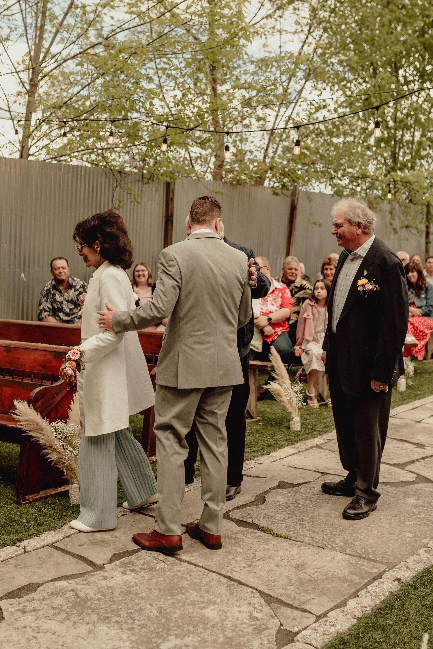 Wedding guests gather outdoors on a stone pathway during a casual outdoor celebration.