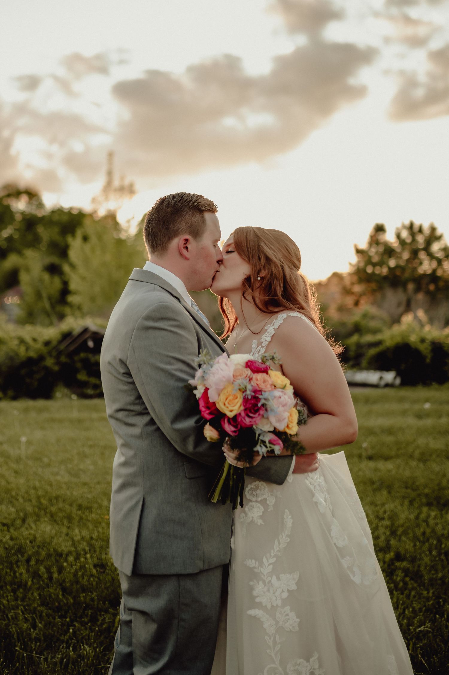 A couple shares a romantic kiss at sunset in a grassy field while holding a vibrant pink and purple bouquet.