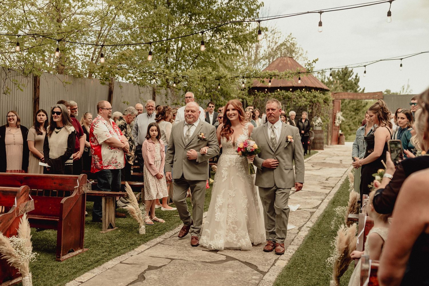 Wedding guests sit outdoors under string lights during a rustic garden celebration at sunset.