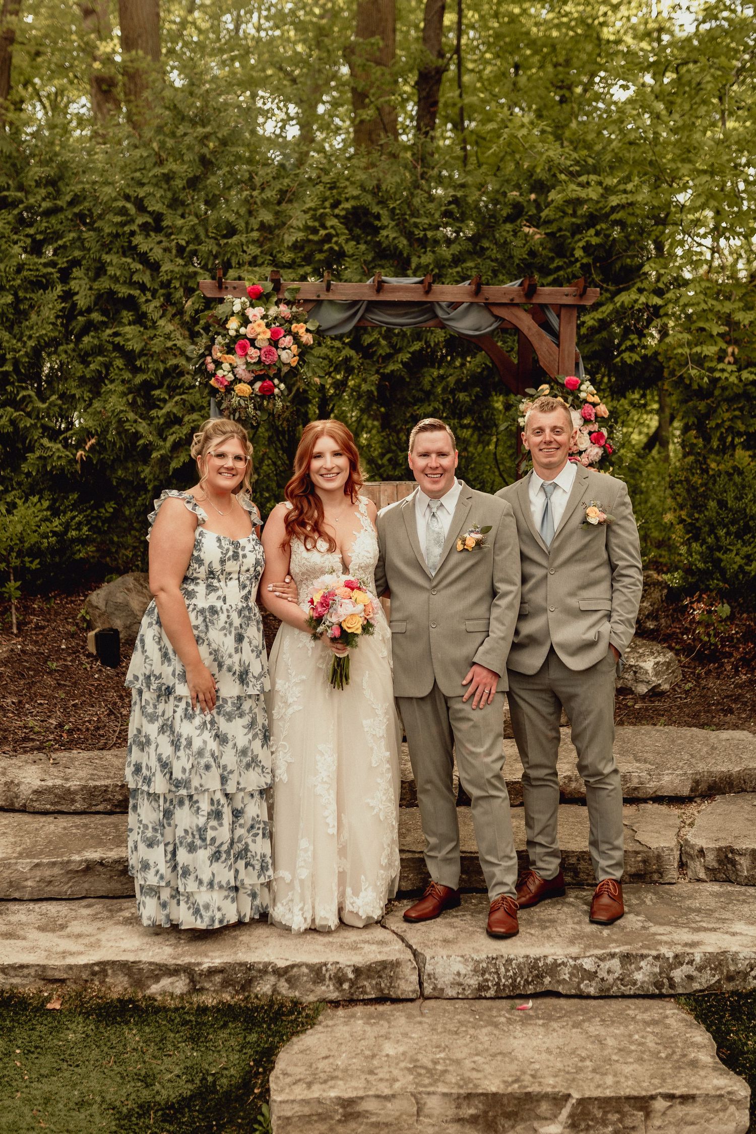 Wedding party members stand together on stone steps in a garden setting wearing gray suits and floral dresses.