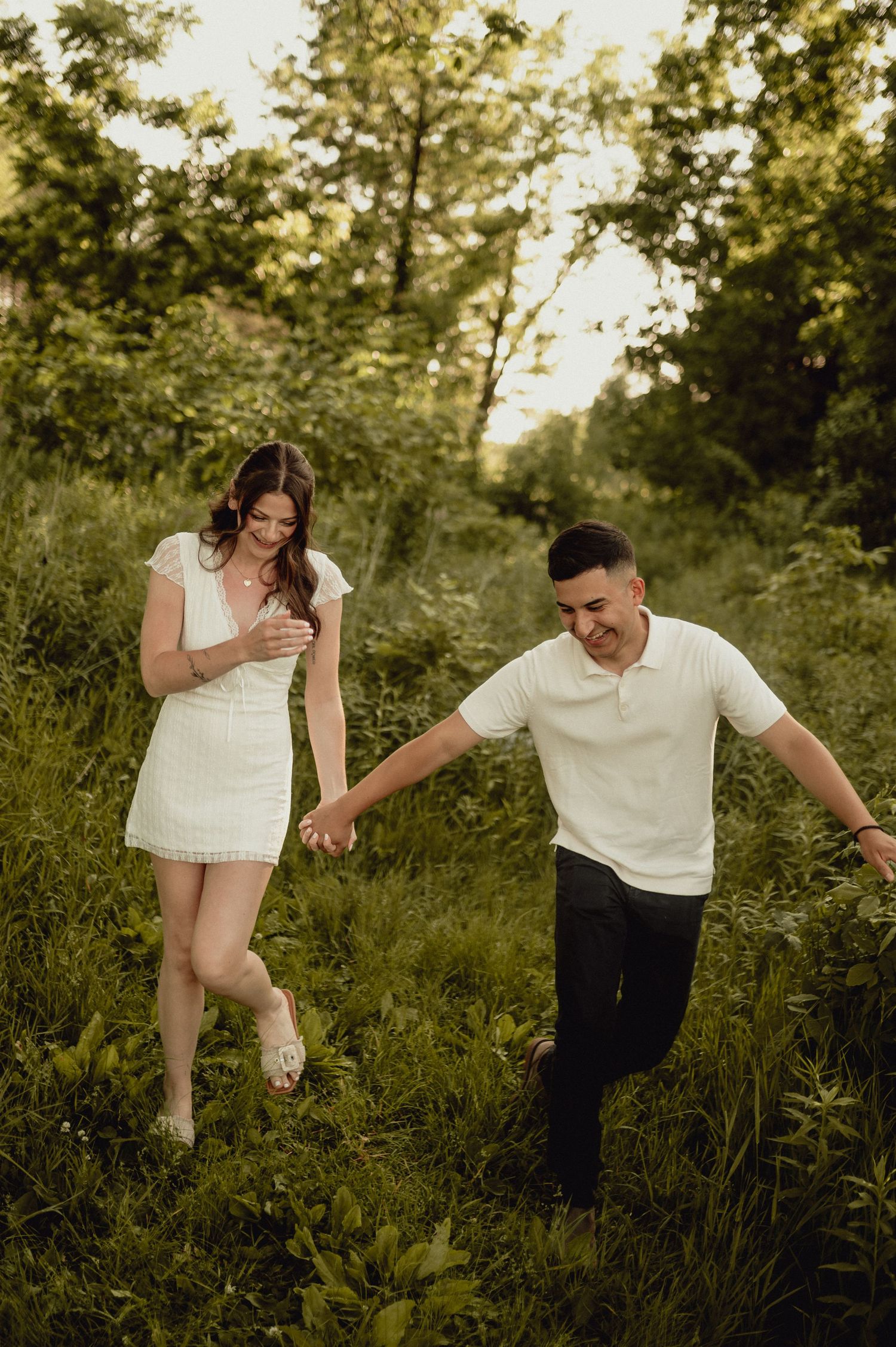 A playful couple holding hands while walking through tall grass in a sunlit field.