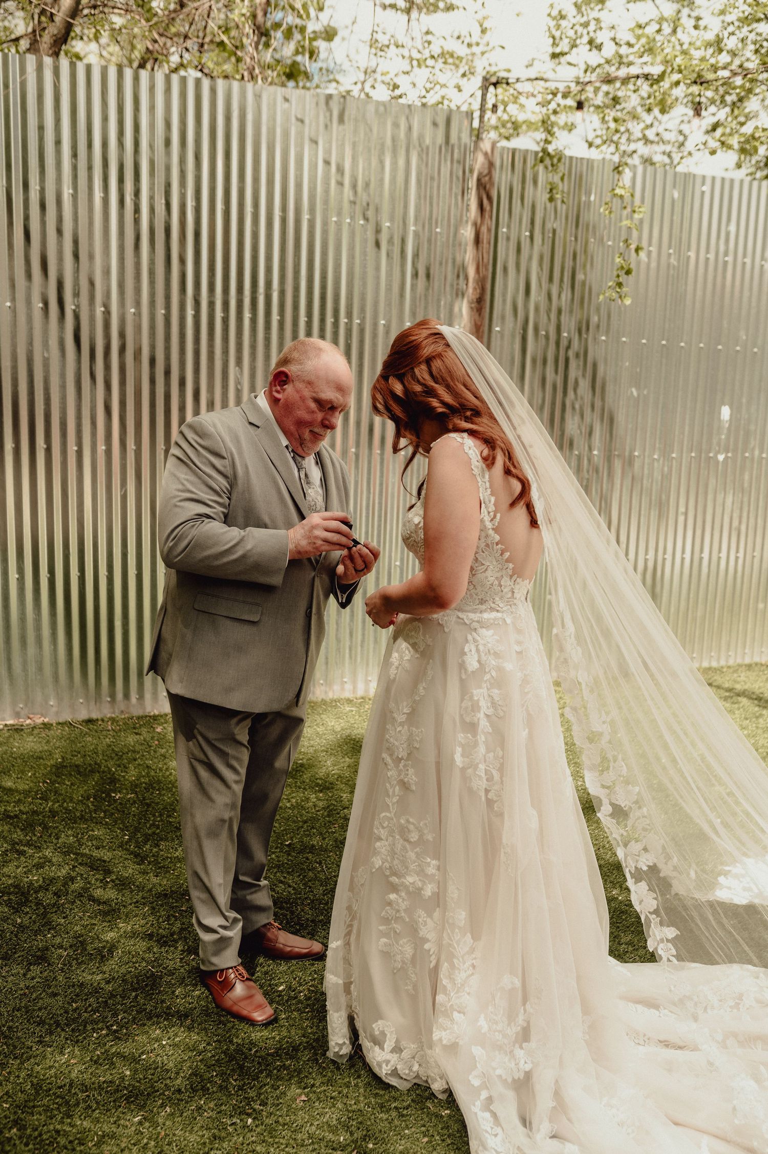 A tender moment at an outdoor wedding as a bride in a lace gown with flowing veil stands on grass near a metal wall.