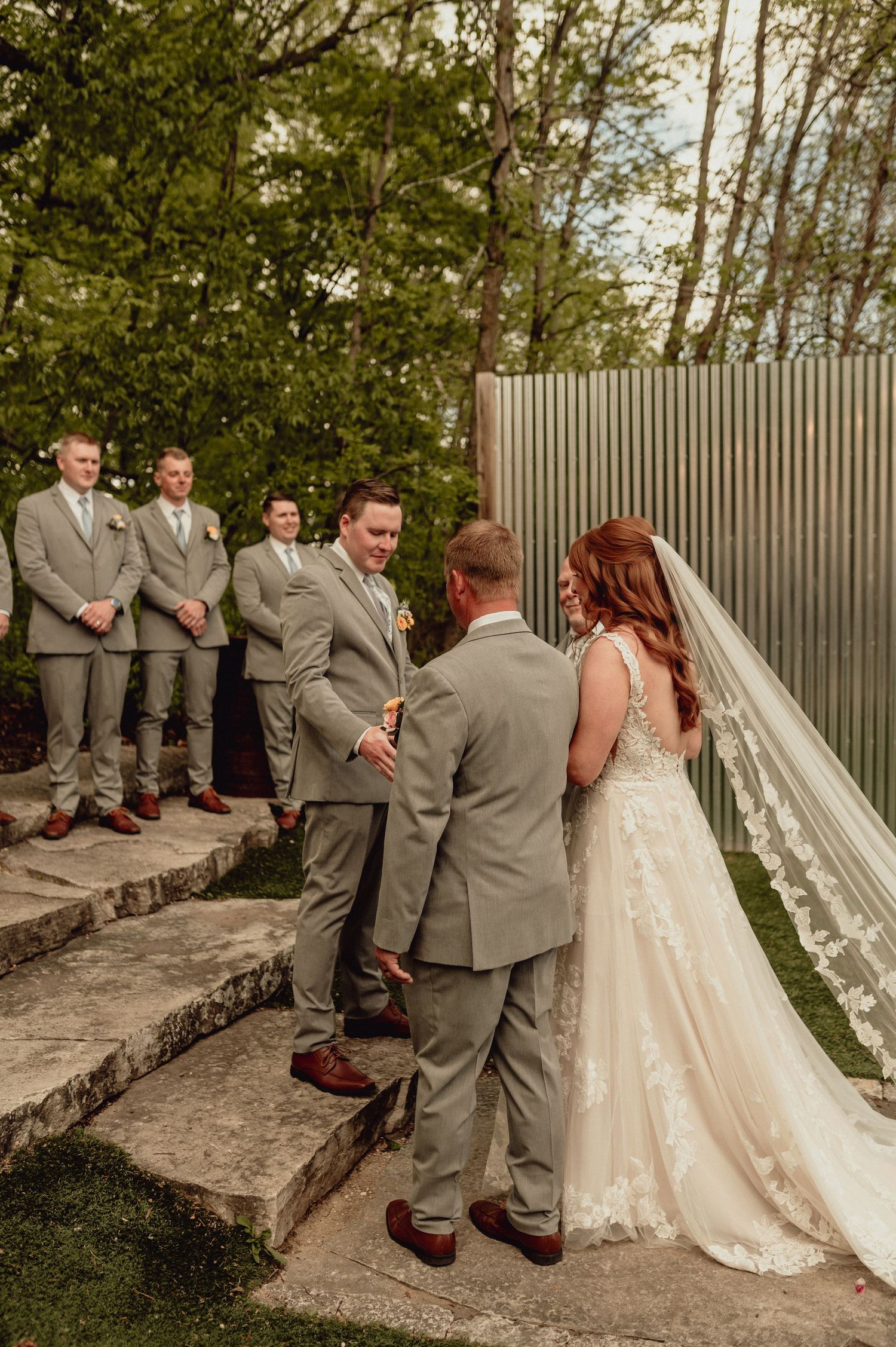 Wedding party in gray suits and a flowing white dress gather for photos on stone steps in an outdoor garden setting.
