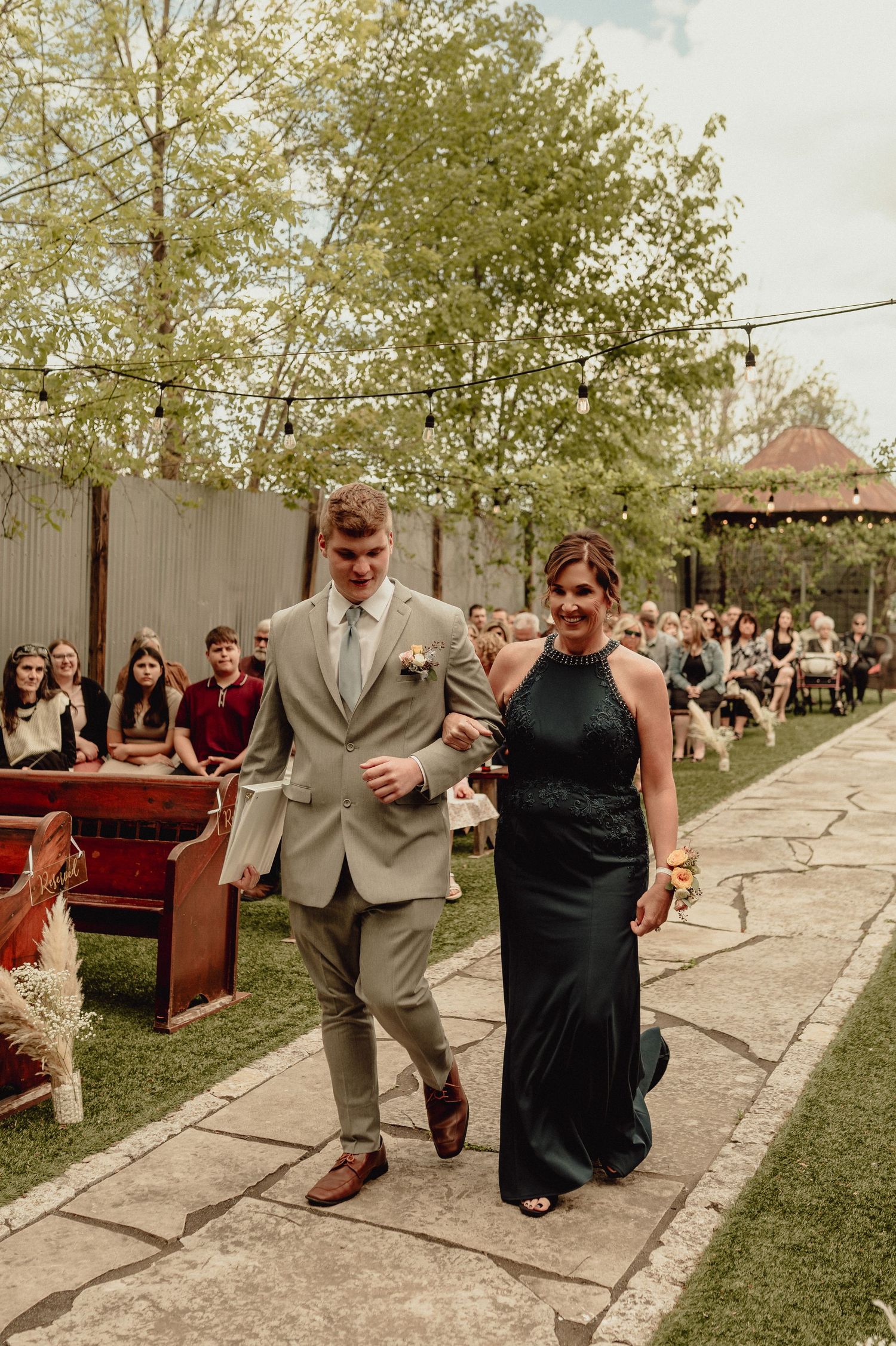 Wedding guests walking down a stone pathway at an outdoor venue with trees and rustic decor in the background.