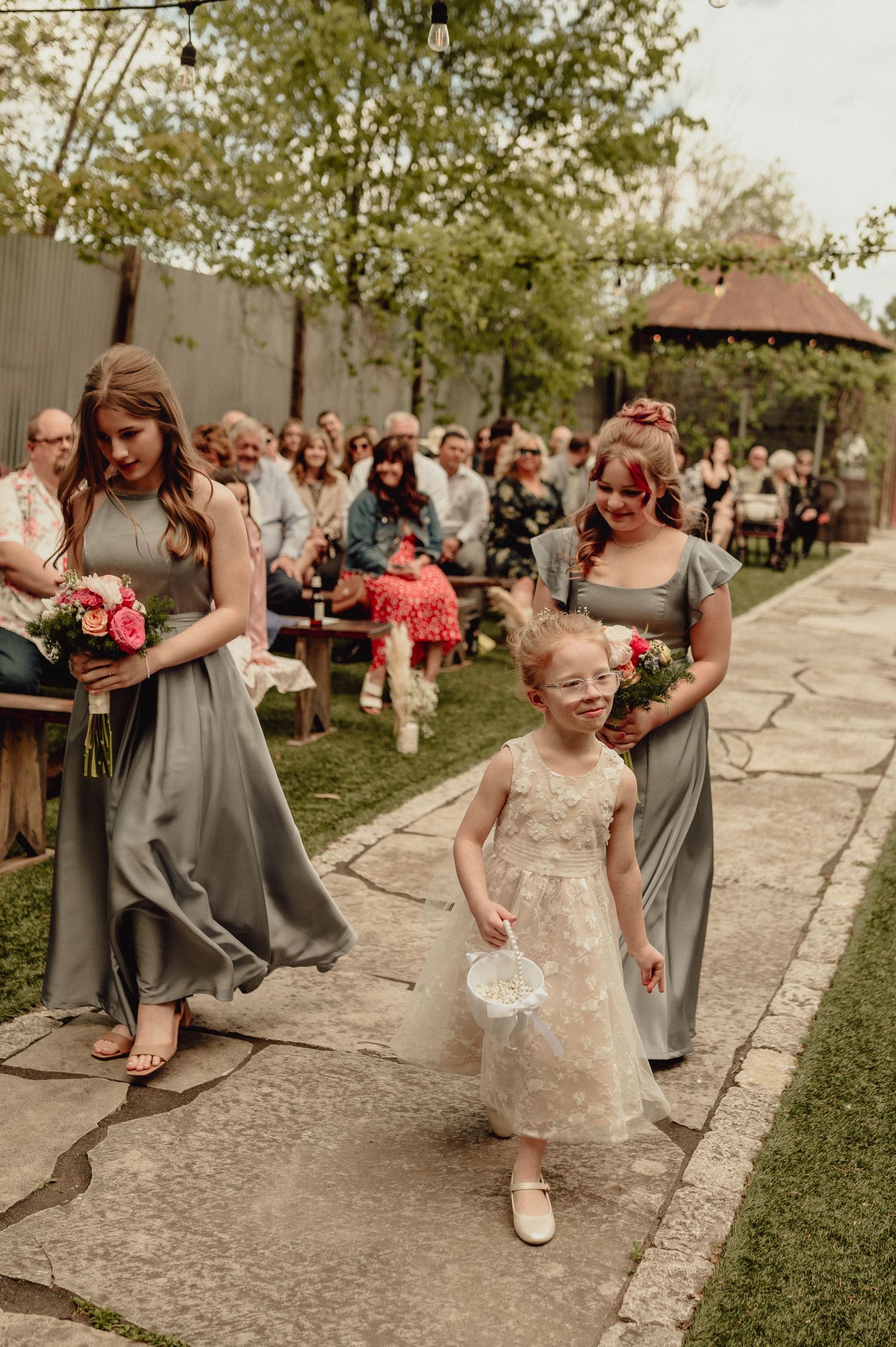 Wedding party members playfully dance and pose together on a stone pathway surrounded by grass and trees.