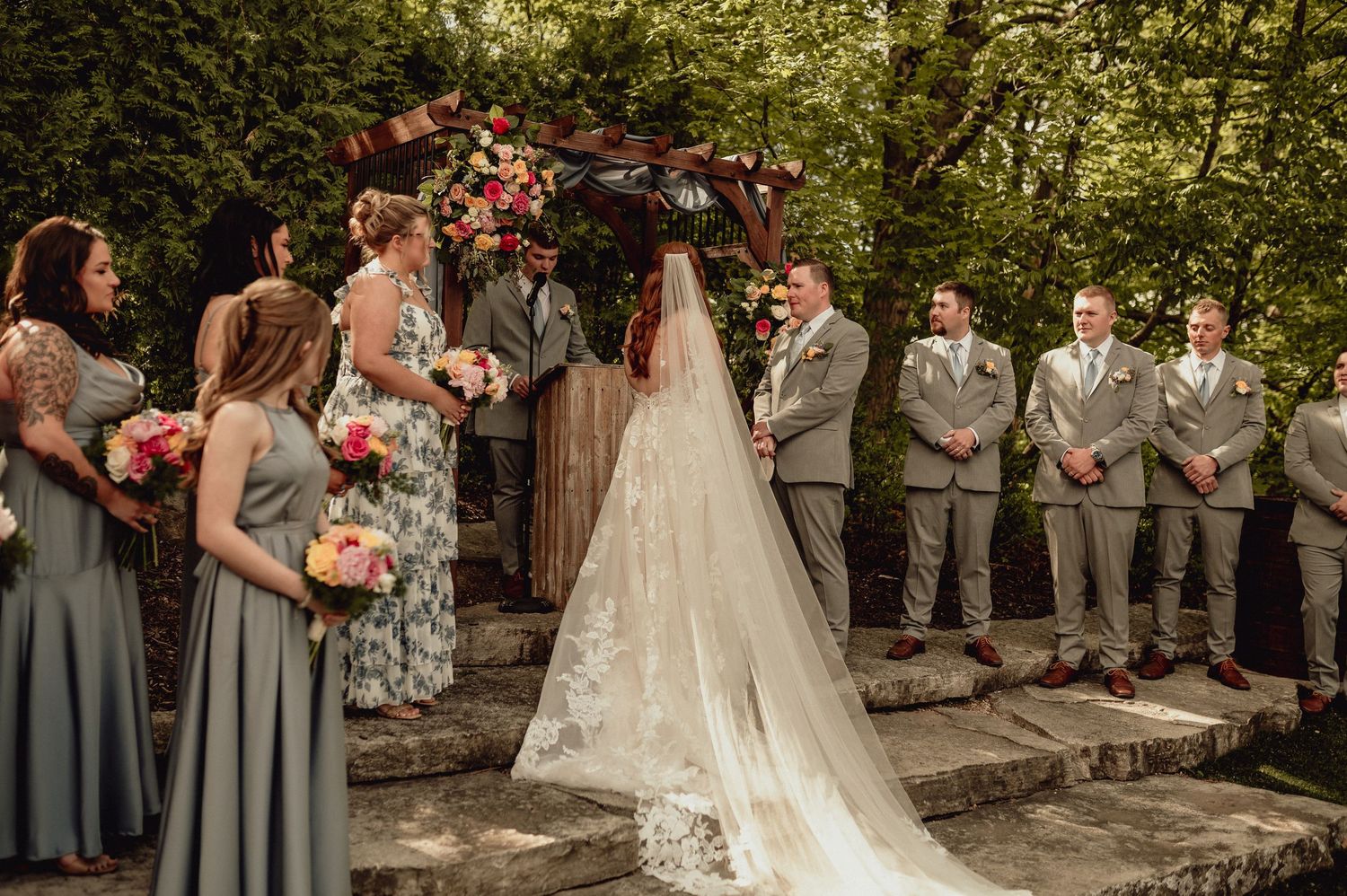 An outdoor wedding ceremony takes place under a floral arch with bridesmaids in sage dresses and groomsmen in gray suits.