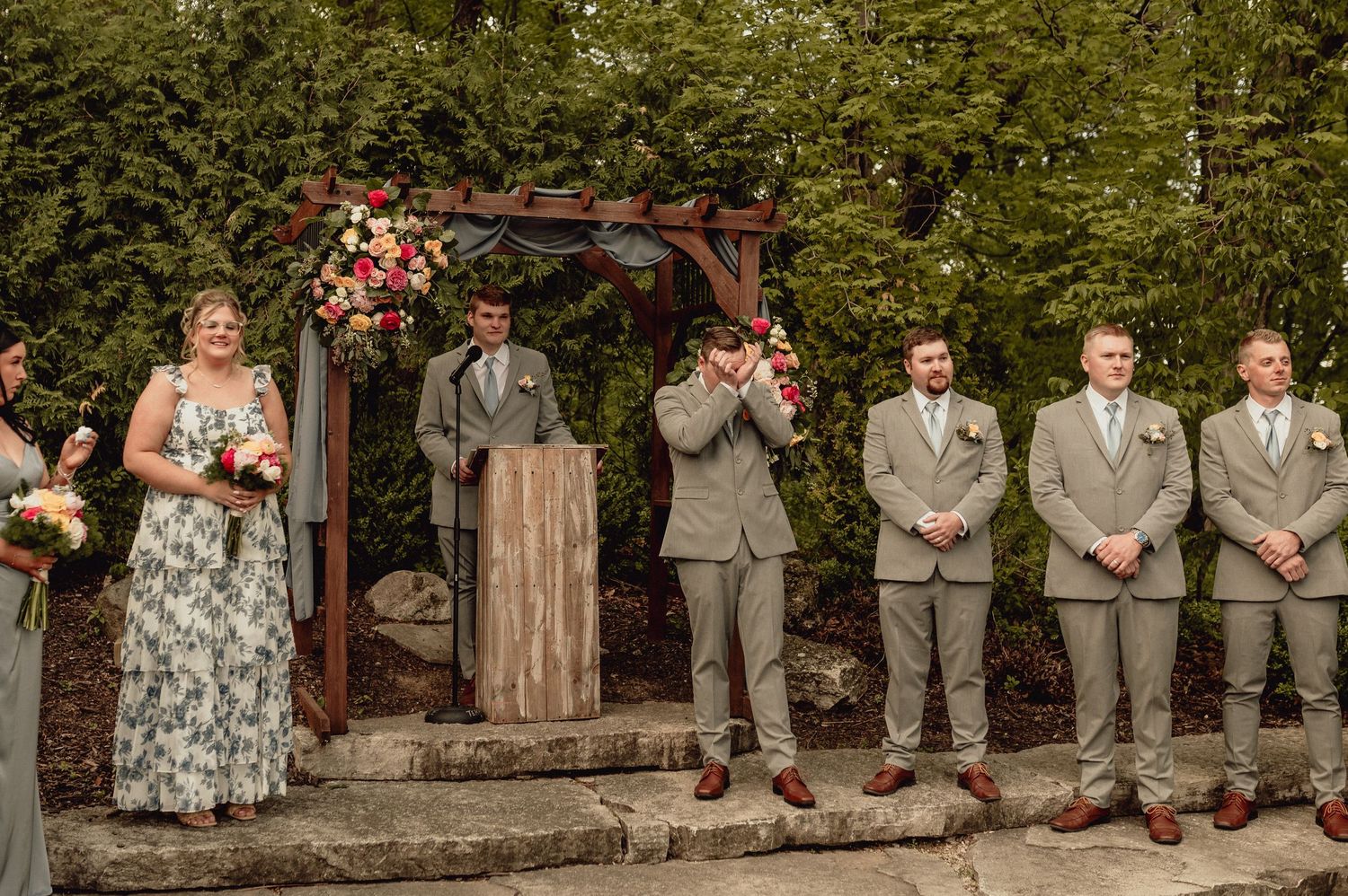 A wedding party poses in a line against a rustic wooden backdrop with lush greenery in the background.