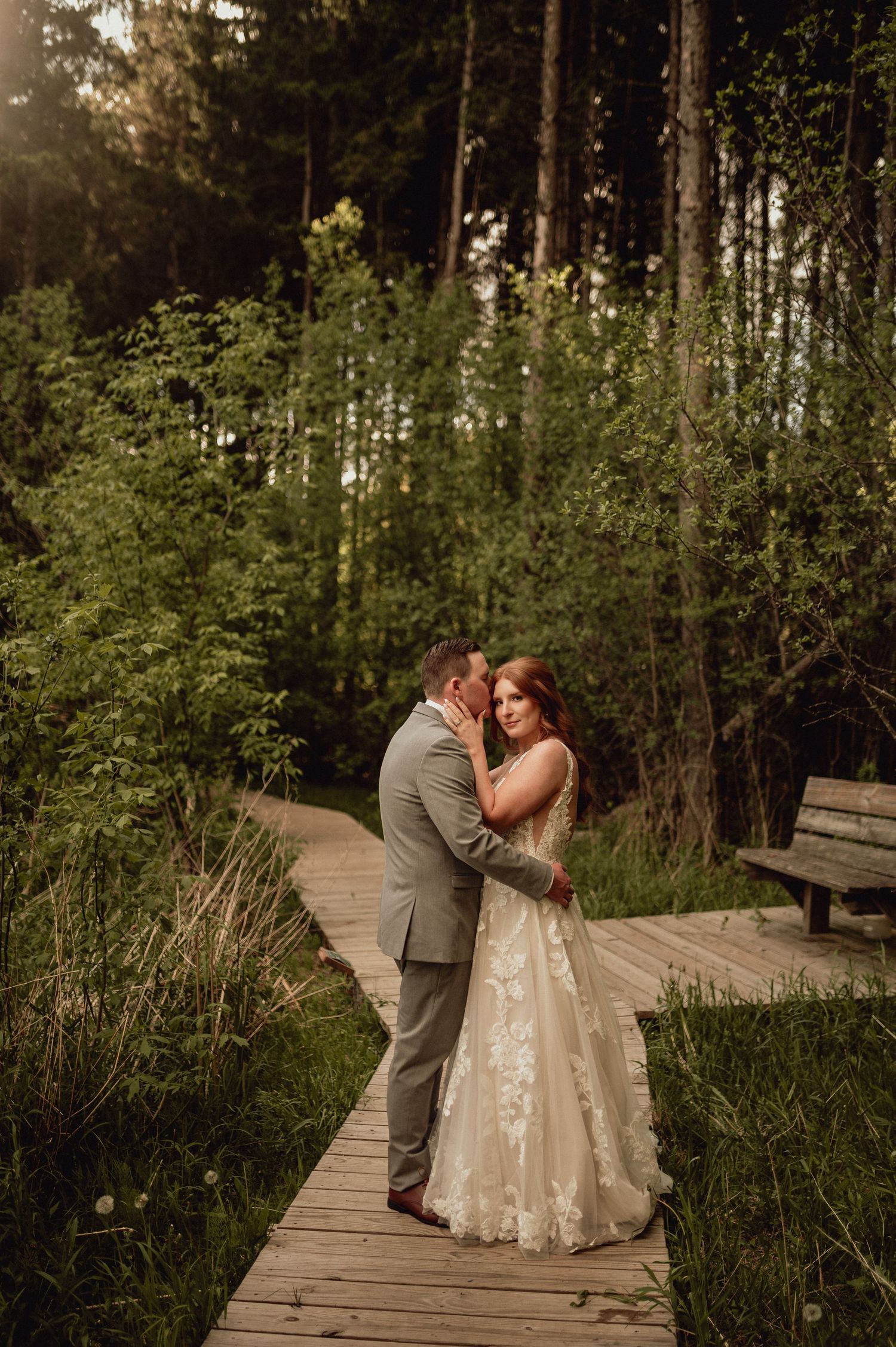 A couple embraces on a wooden boardwalk surrounded by tall trees and natural greenery at sunset.