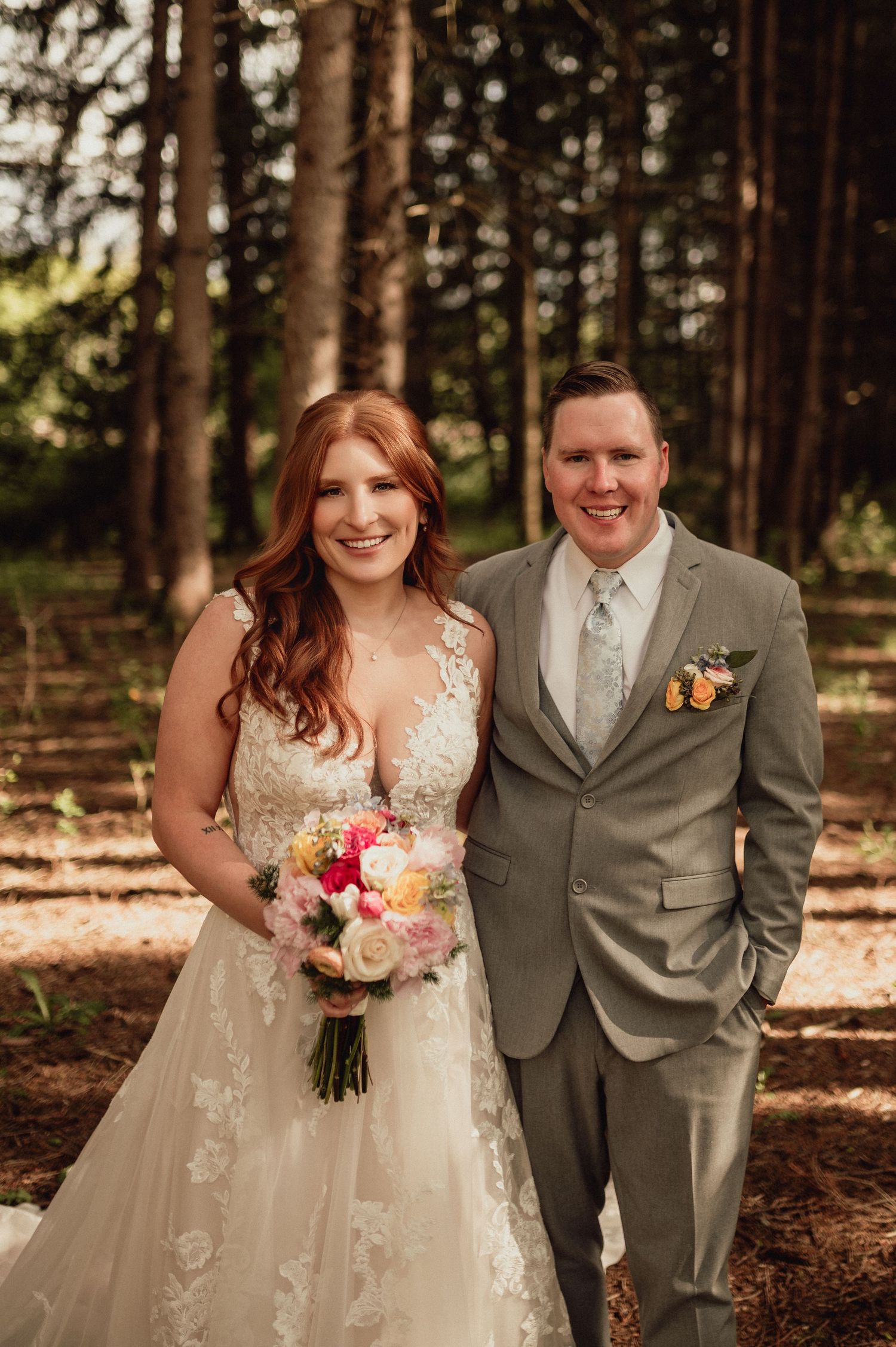 A wedding couple poses in a forest setting with the bride in a lace gown holding a pink and peach bouquet.