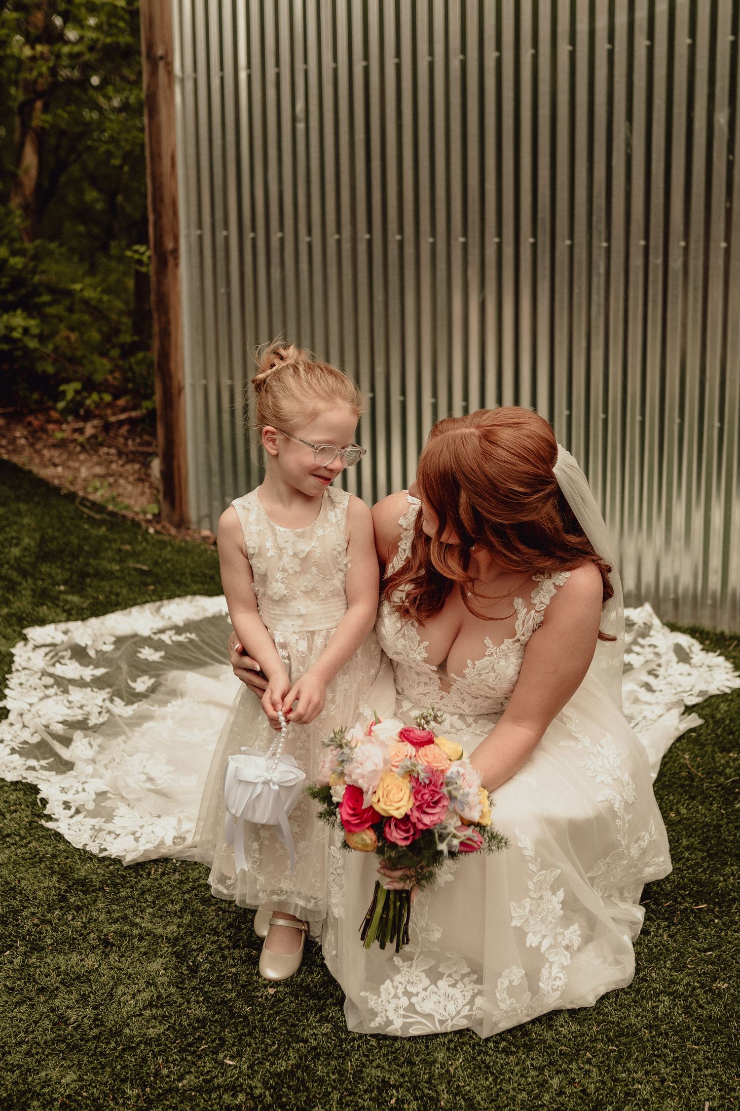 A bride and young girl in matching white lace dresses sit together on grass with a colorful floral bouquet.