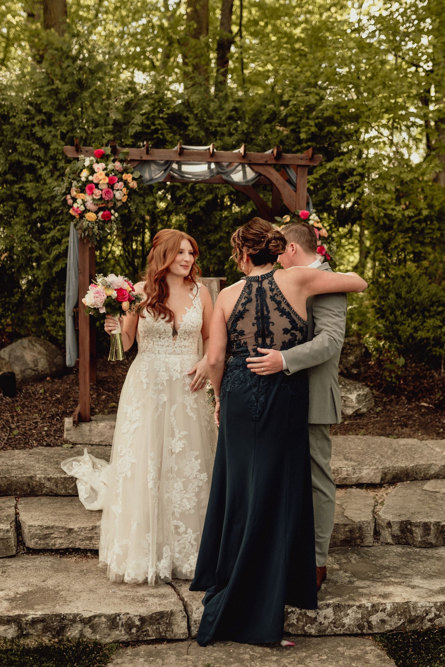 Two people share a joyful moment in front of a wooden arch decorated with pink flowers at an outdoor wedding ceremony.