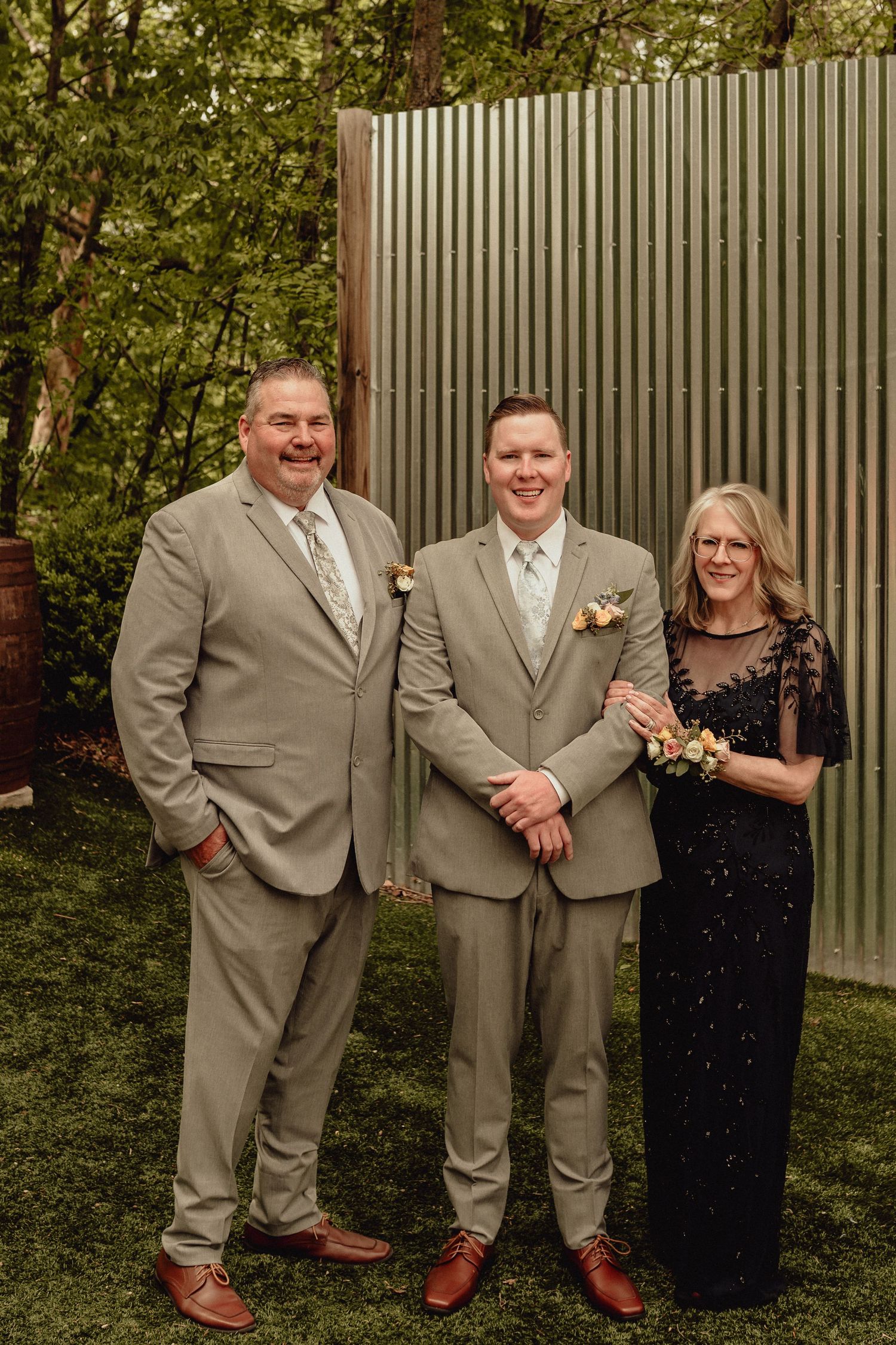 Three people in formal attire stand together outdoors against a fence and greenery backdrop.