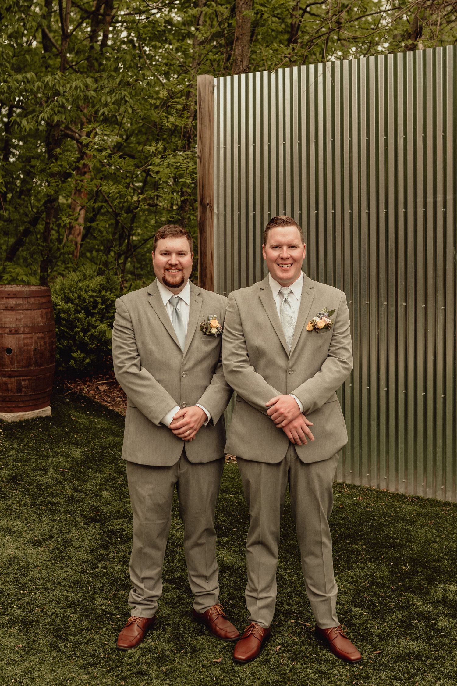 Two groomsmen in light gray suits and brown shoes stand together on grass against a metal fence backdrop.