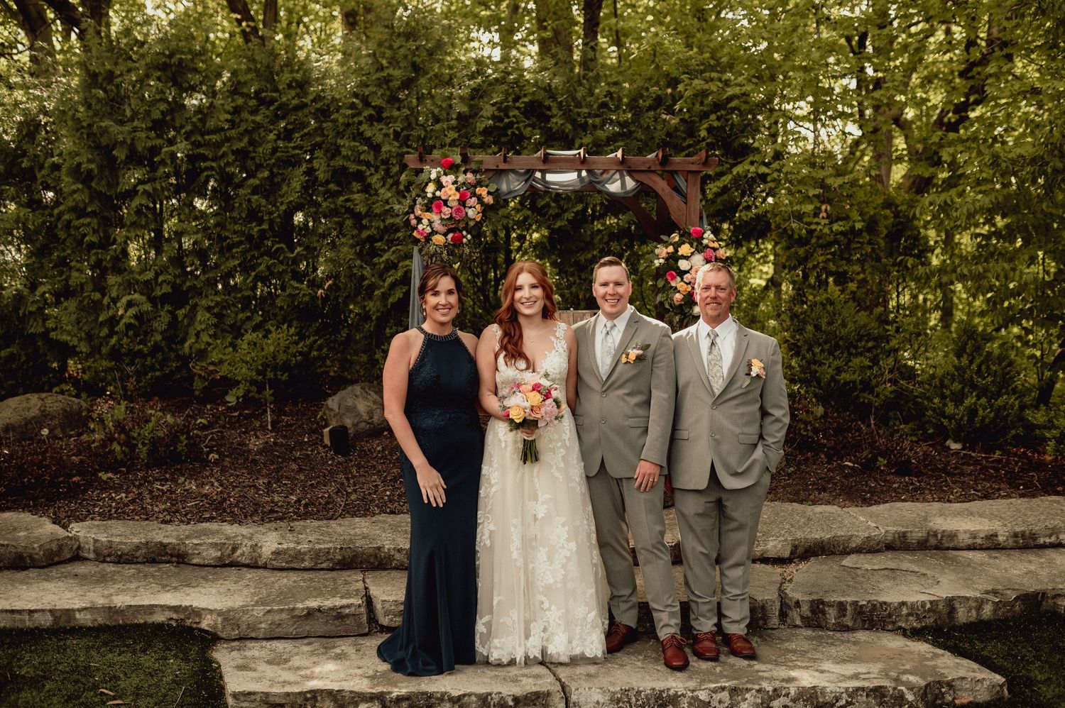 Wedding party poses for outdoor photos in a garden setting with stone walls and lush greenery.