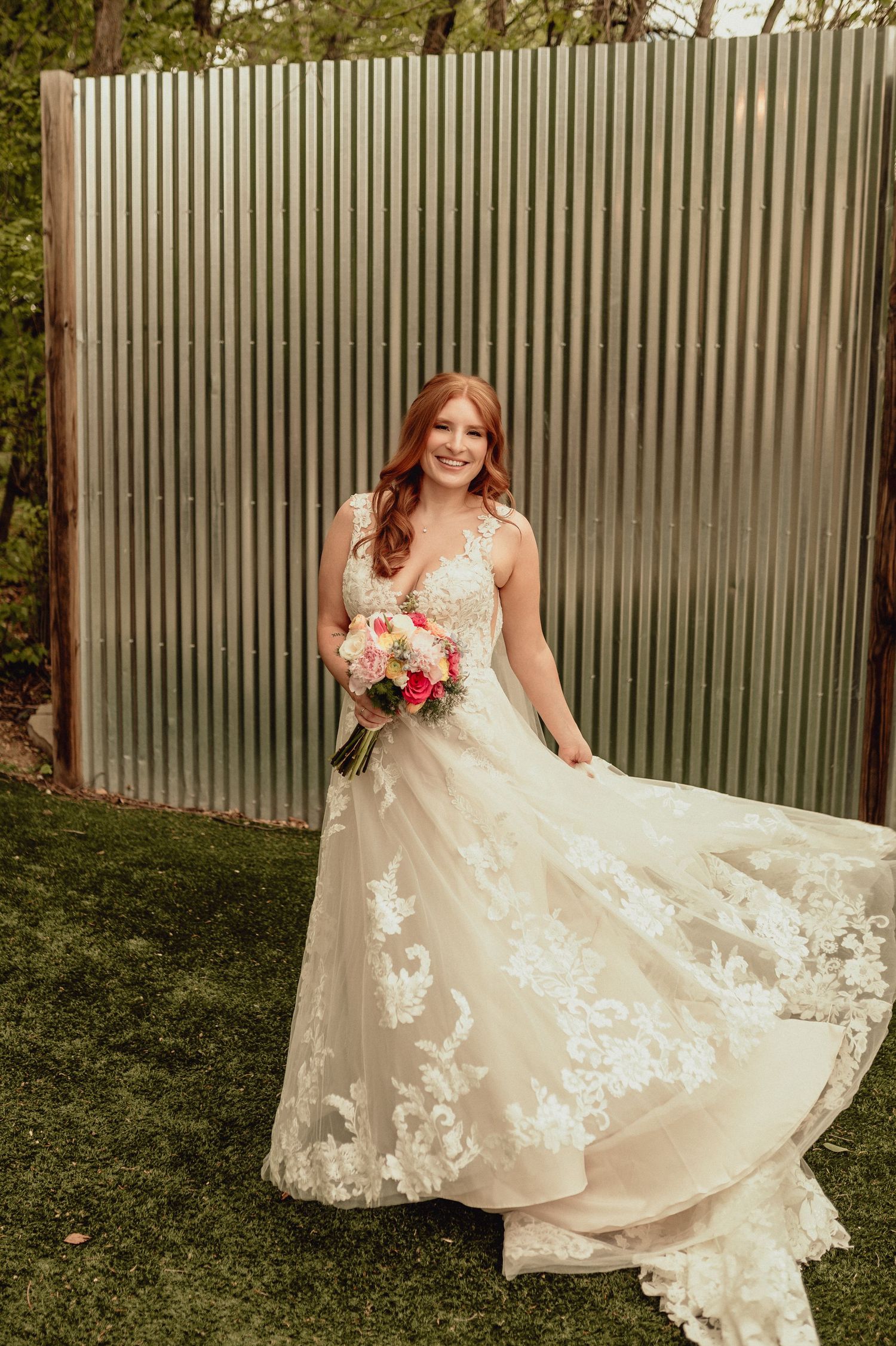 A bride in a flowing lace wedding gown with floral details poses against a corrugated metal wall while holding a bouquet.