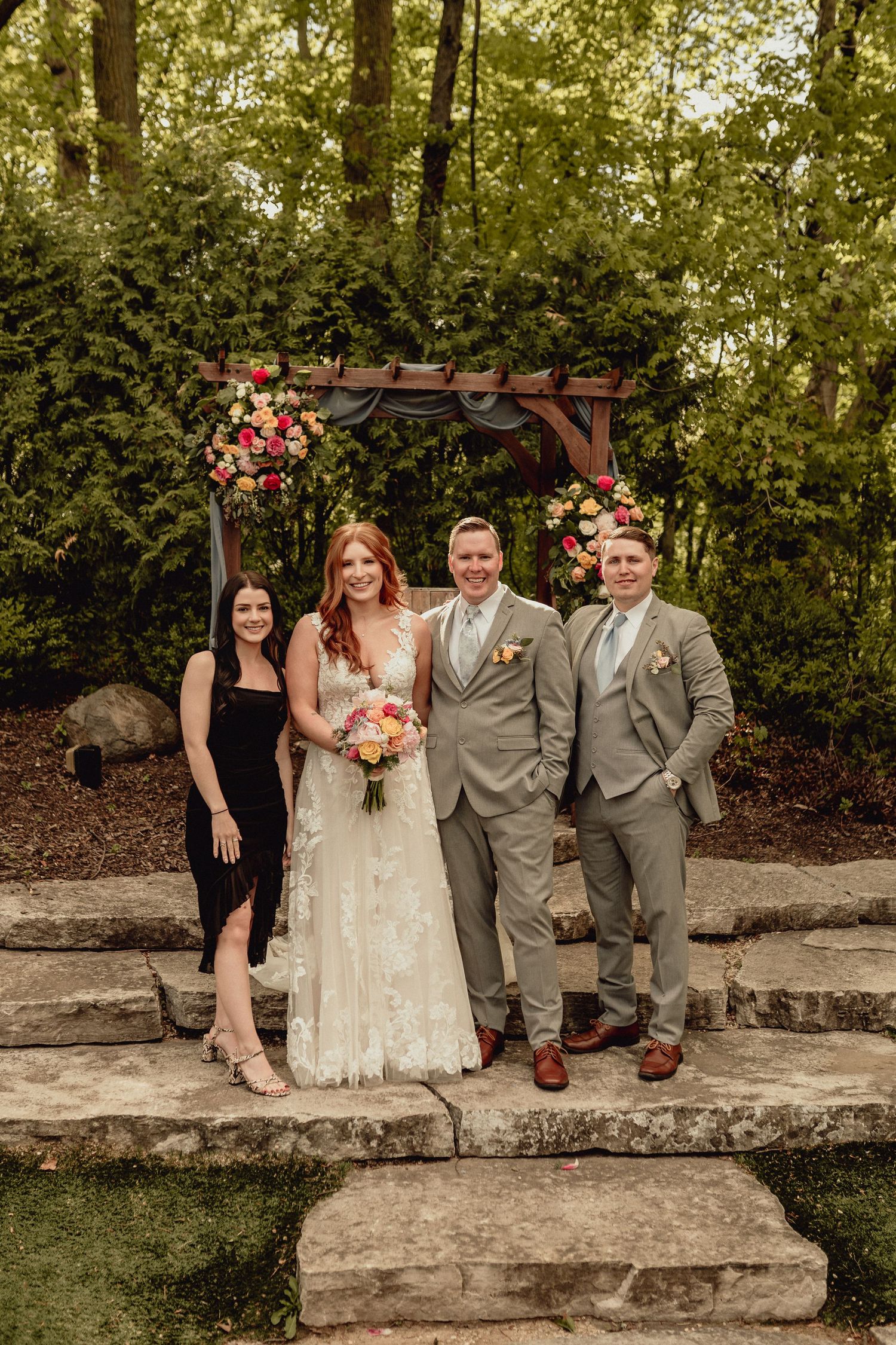 Wedding party poses together under a wooden arbor decorated with pink flowers in an outdoor garden setting.