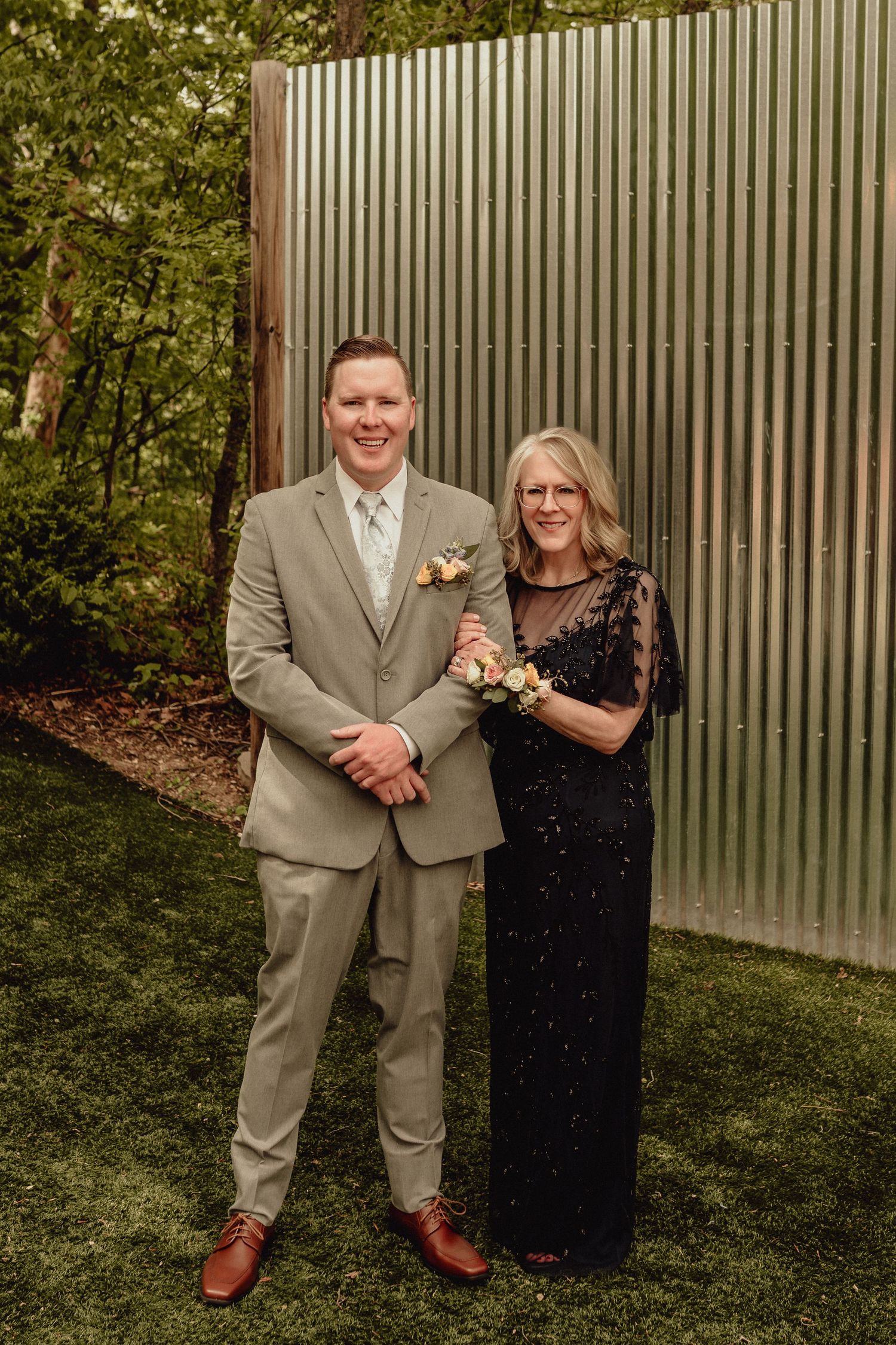 A couple poses together outdoors against a metal wall, wearing formal attire with the person in a light suit and brown shoes.