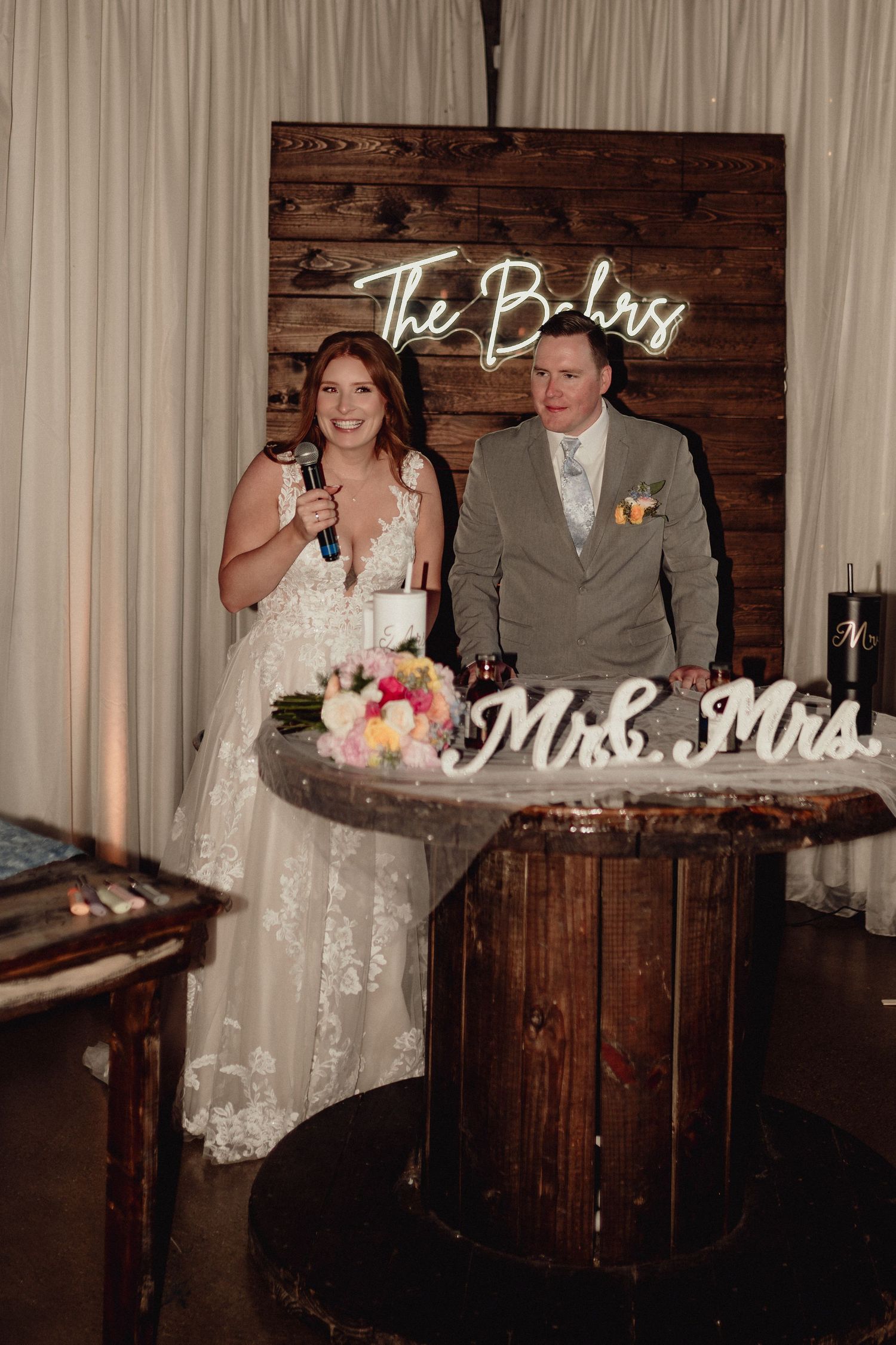 Newlyweds give speeches at rustic wooden bar during wedding reception with Mr & Mrs sign visible.