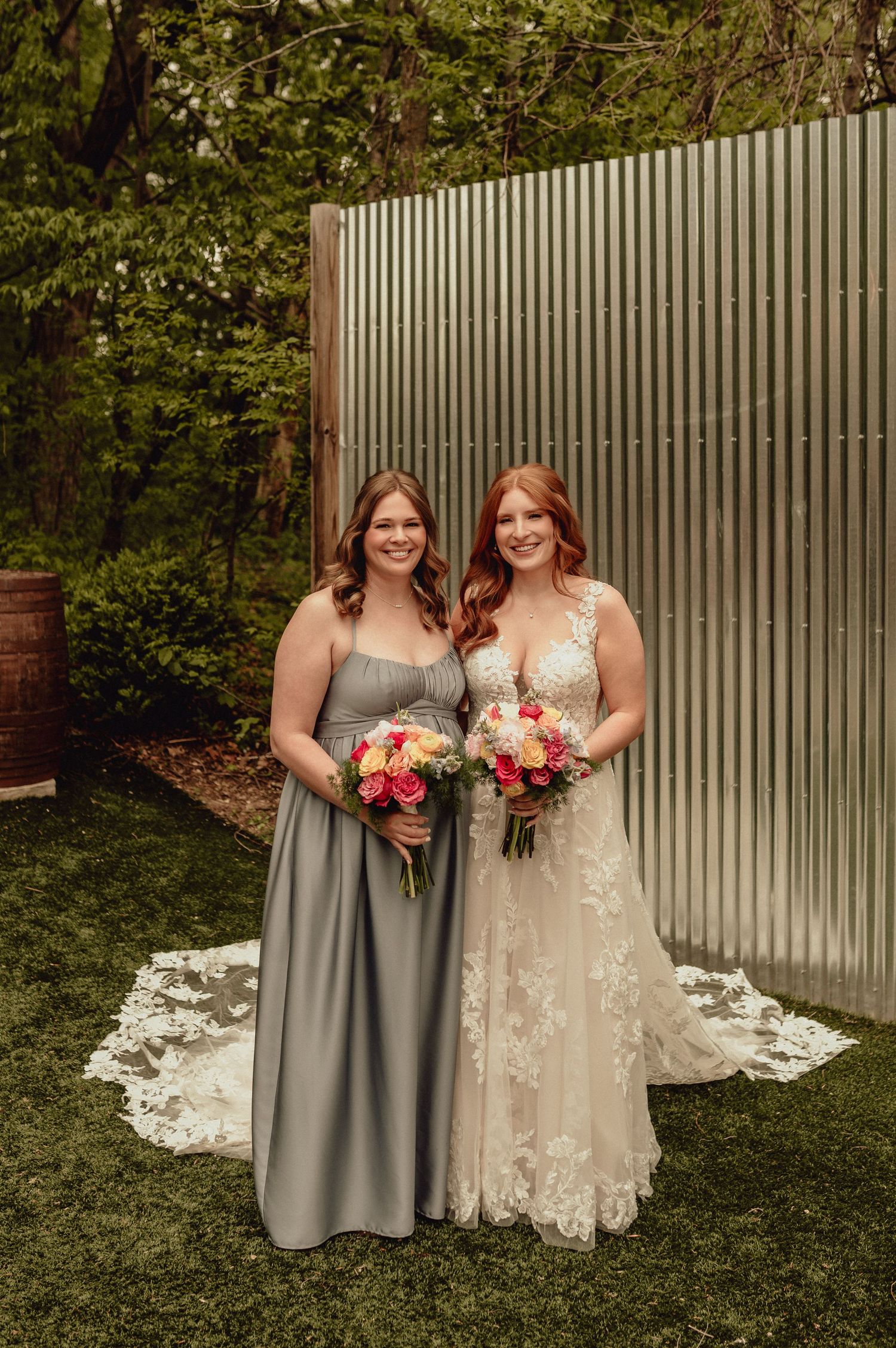 Two people in wedding attire pose together outdoors against a white fence with pink floral bouquets.