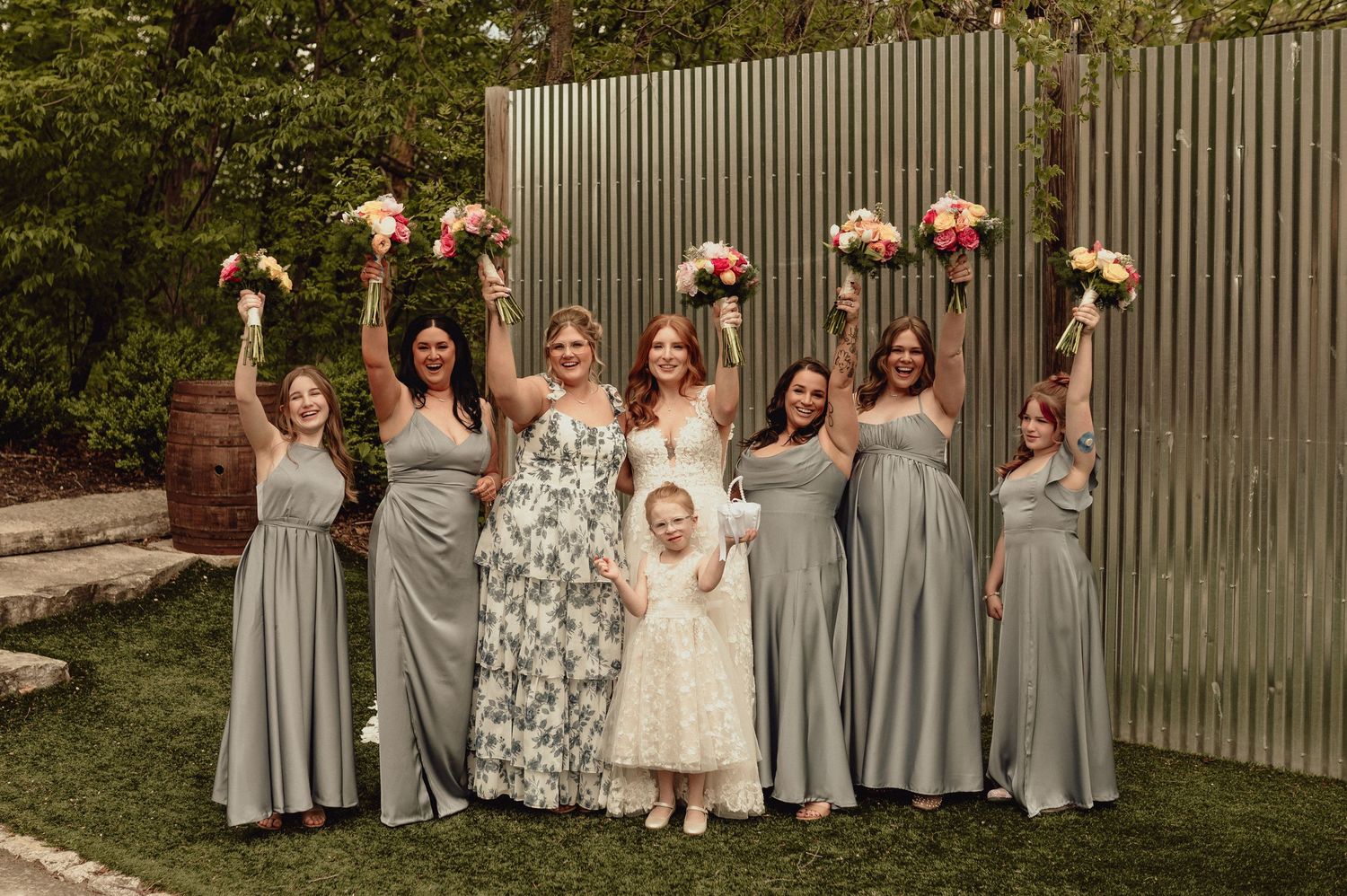 Wedding party in gray bridesmaid dresses and white bridal gown pose together in outdoor garden setting.