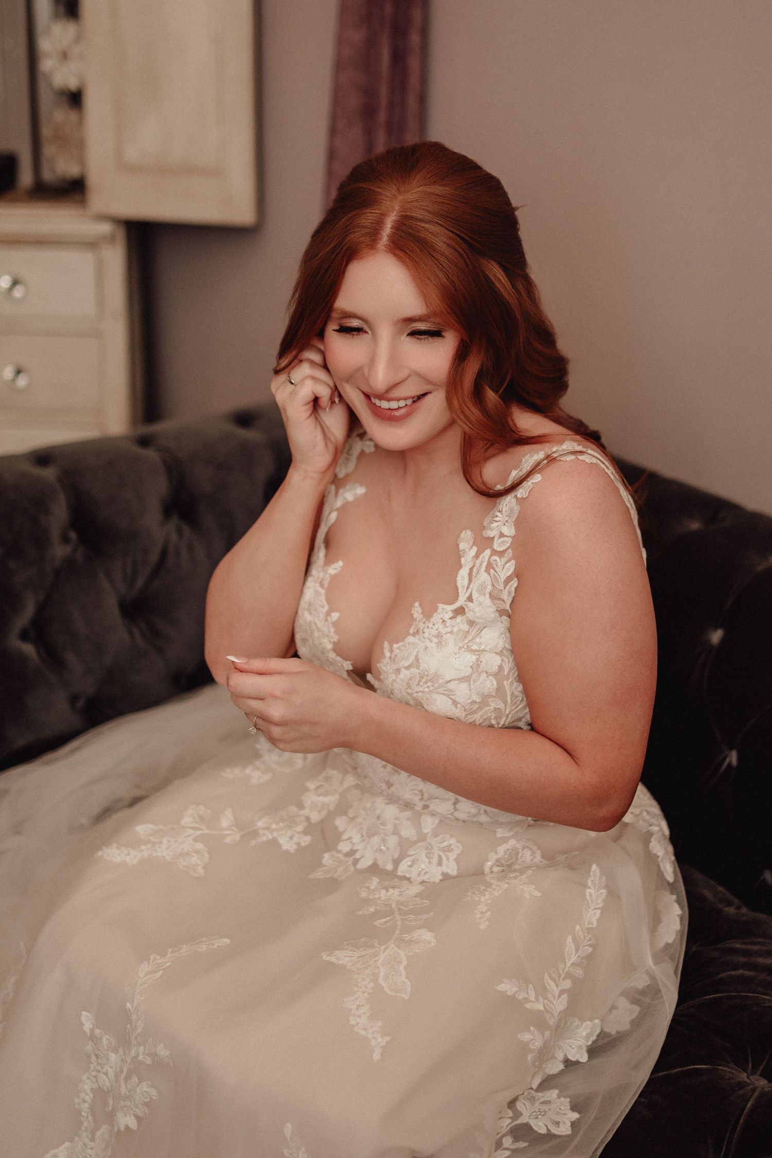 A bride in a white lace wedding dress sits on a dark couch while laughing and looking down.