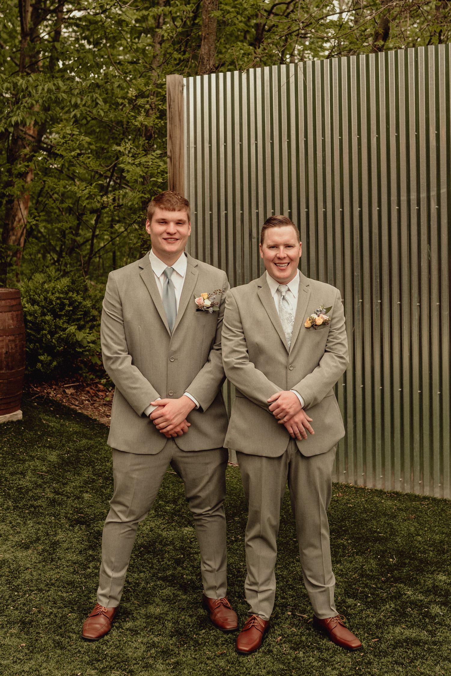 Two people stand together wearing matching tan suits and brown shoes on a grassy lawn with a fence behind them.