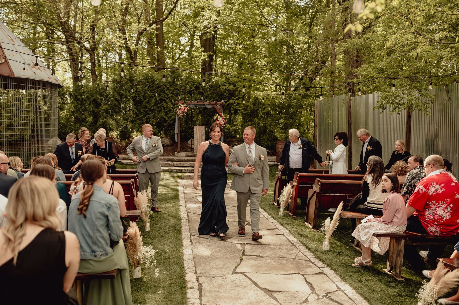 Wedding guests gather outdoors beneath trees during a summer garden ceremony celebration.