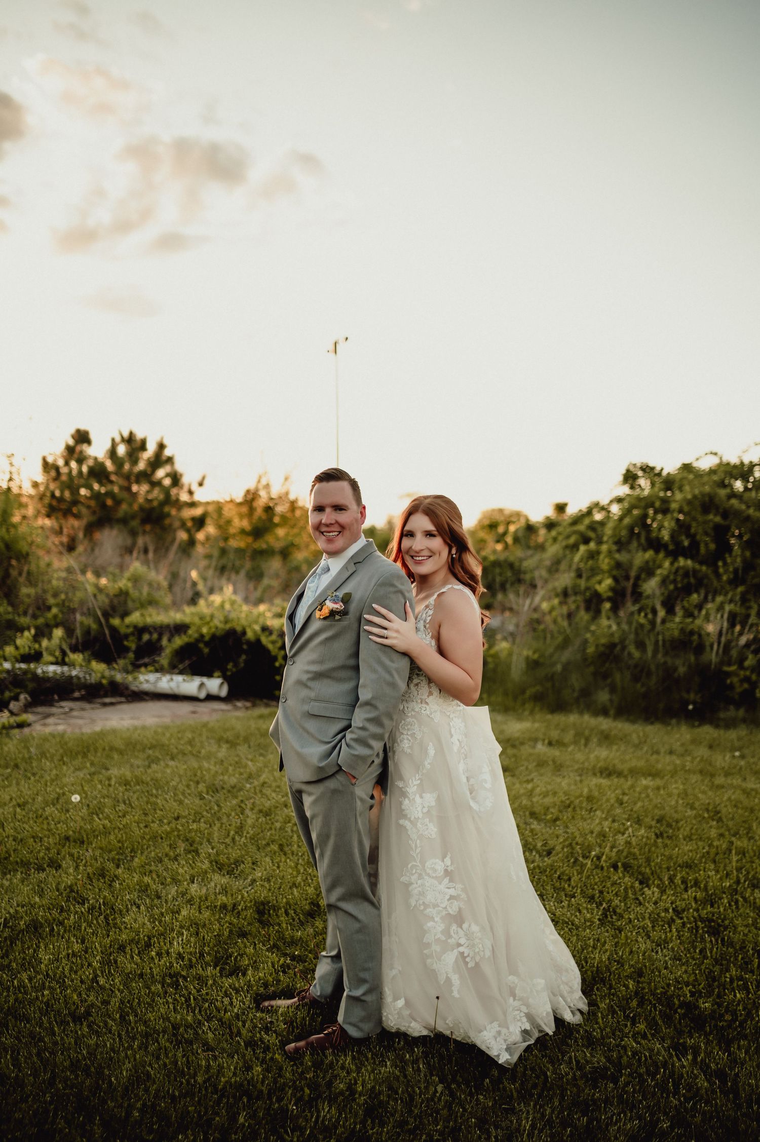A couple in wedding attire embrace while standing in a grassy field during golden hour.