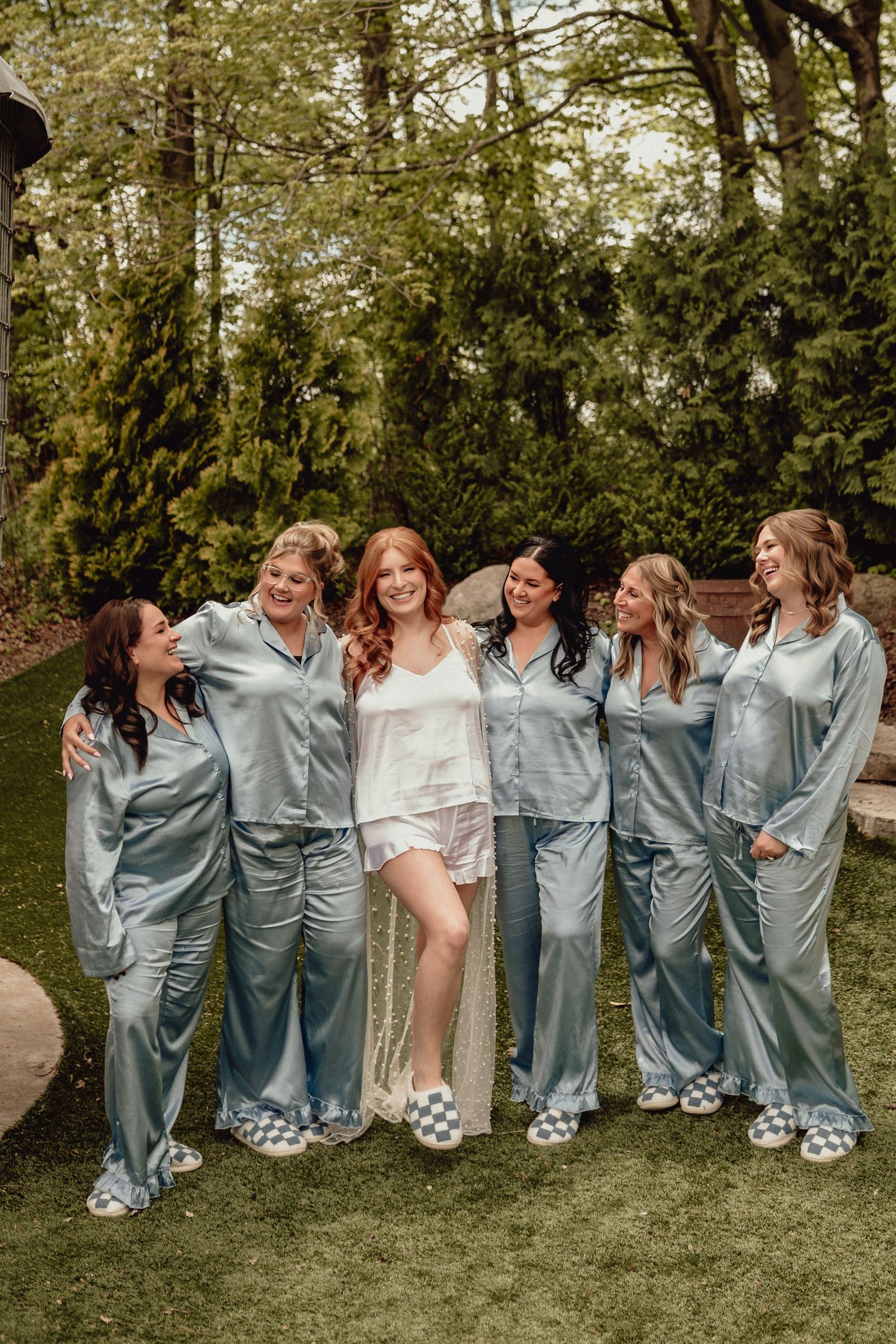 A group wearing matching light blue satin pajamas poses playfully on grass during a wedding celebration.