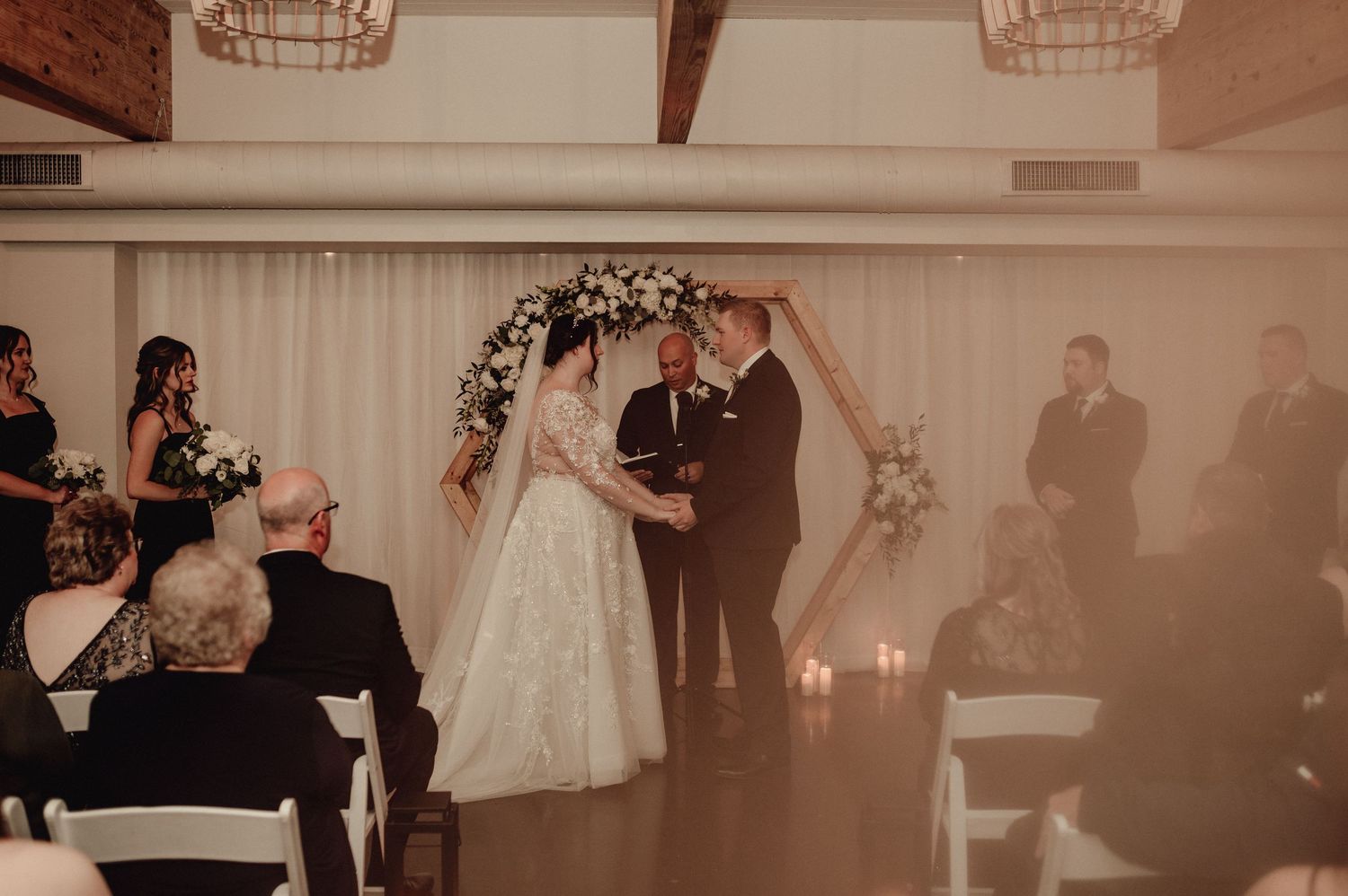A wedding reception venue decorated with white curtains and floral arrangements shows multiple tables and guests in formal attire.
