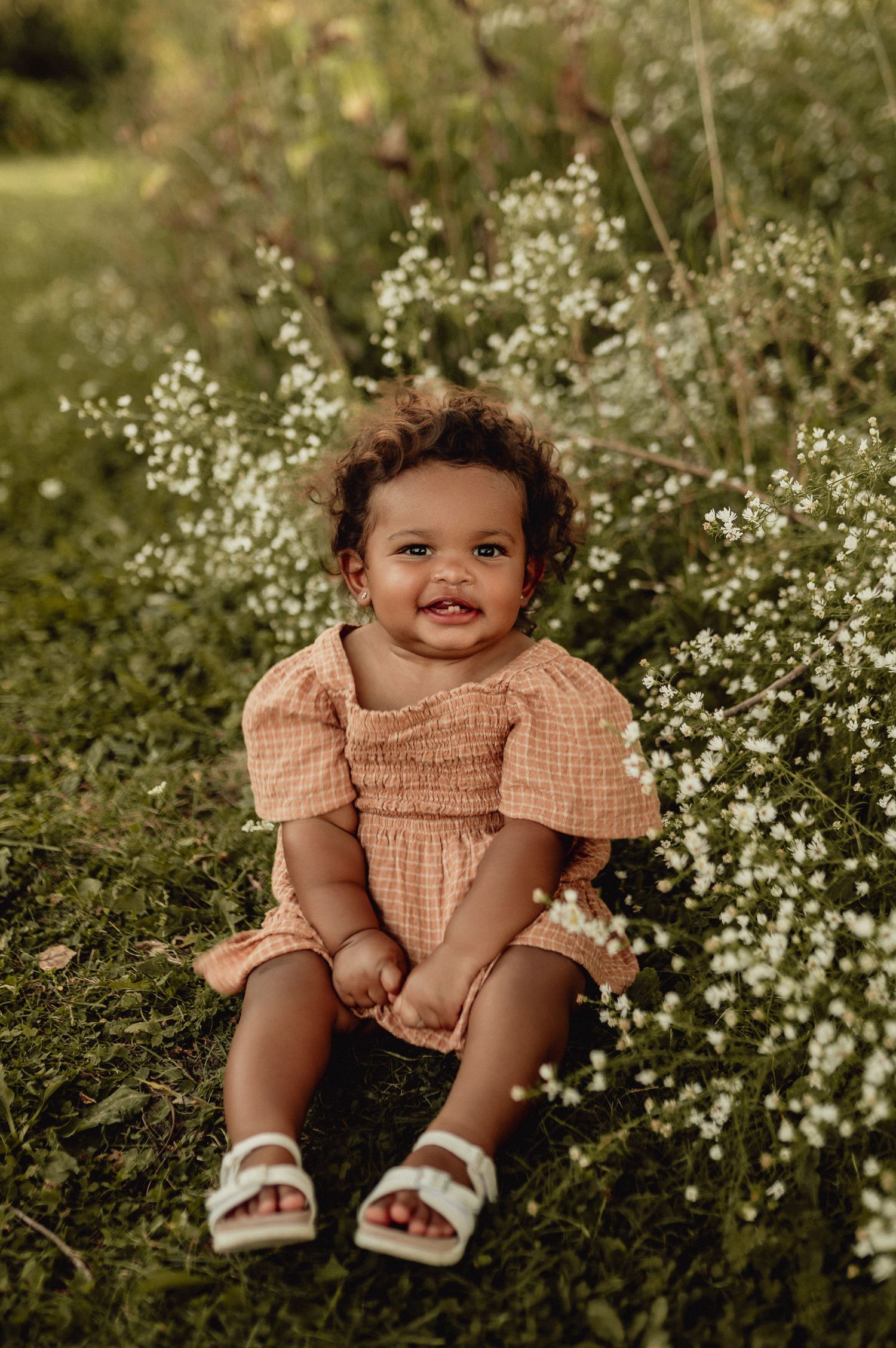 Baby in gingham dress sitting among white wildflowers in garden setting.