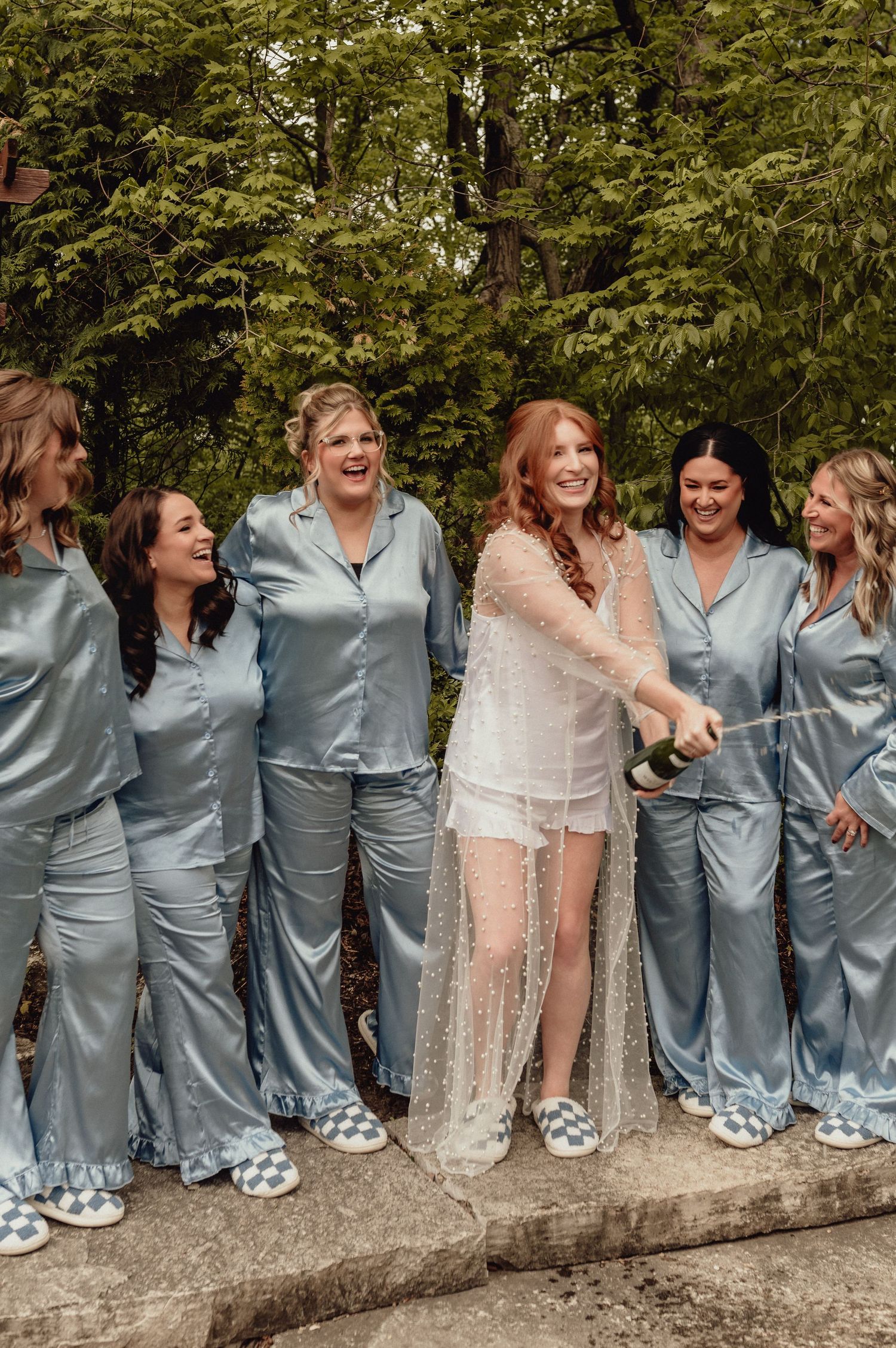Bridal party in matching silk grey pajamas poses together on stone wall outdoors.
