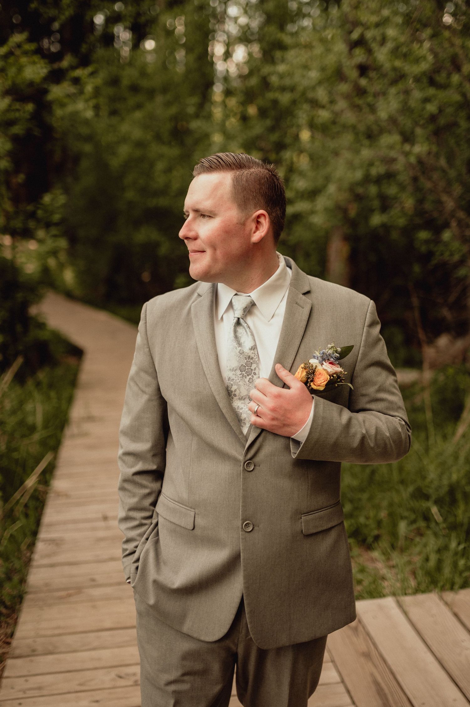 A groom in a light gray suit and tie poses on a wooden boardwalk surrounded by lush greenery.