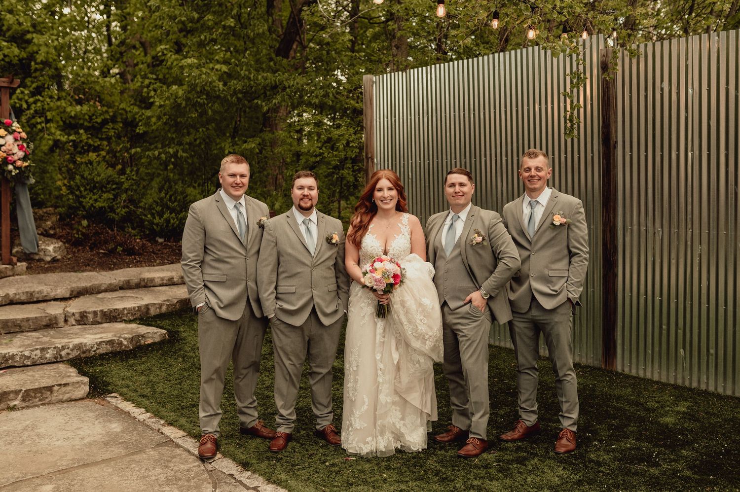 Wedding party in gray suits and pink dresses pose together in front of corrugated metal wall during outdoor celebration.