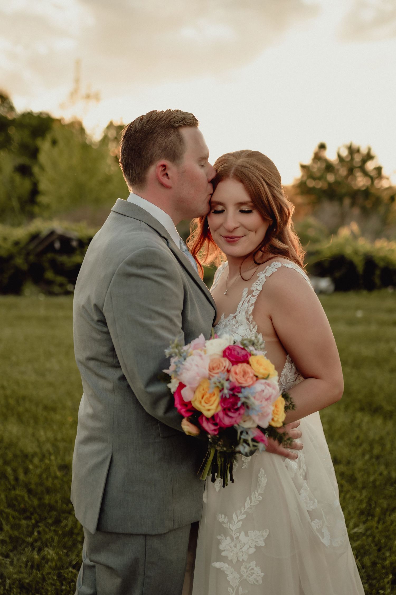 Couple shares tender moment at sunset wedding with vibrant pink and yellow bouquet in grassy outdoor setting.