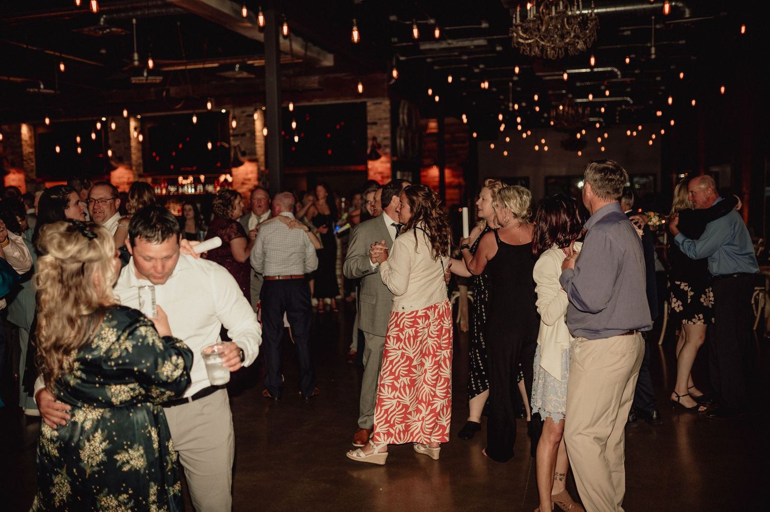 People dancing together at a dimly lit party or celebration event with a chandelier overhead.
