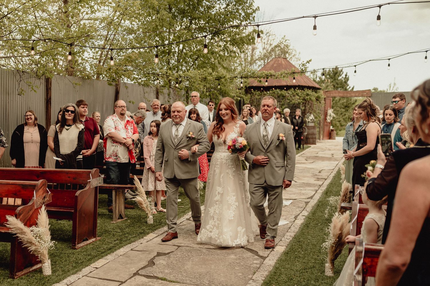 Wedding ceremony guests walk down an outdoor aisle decorated with string lights and natural greenery at a rustic venue.