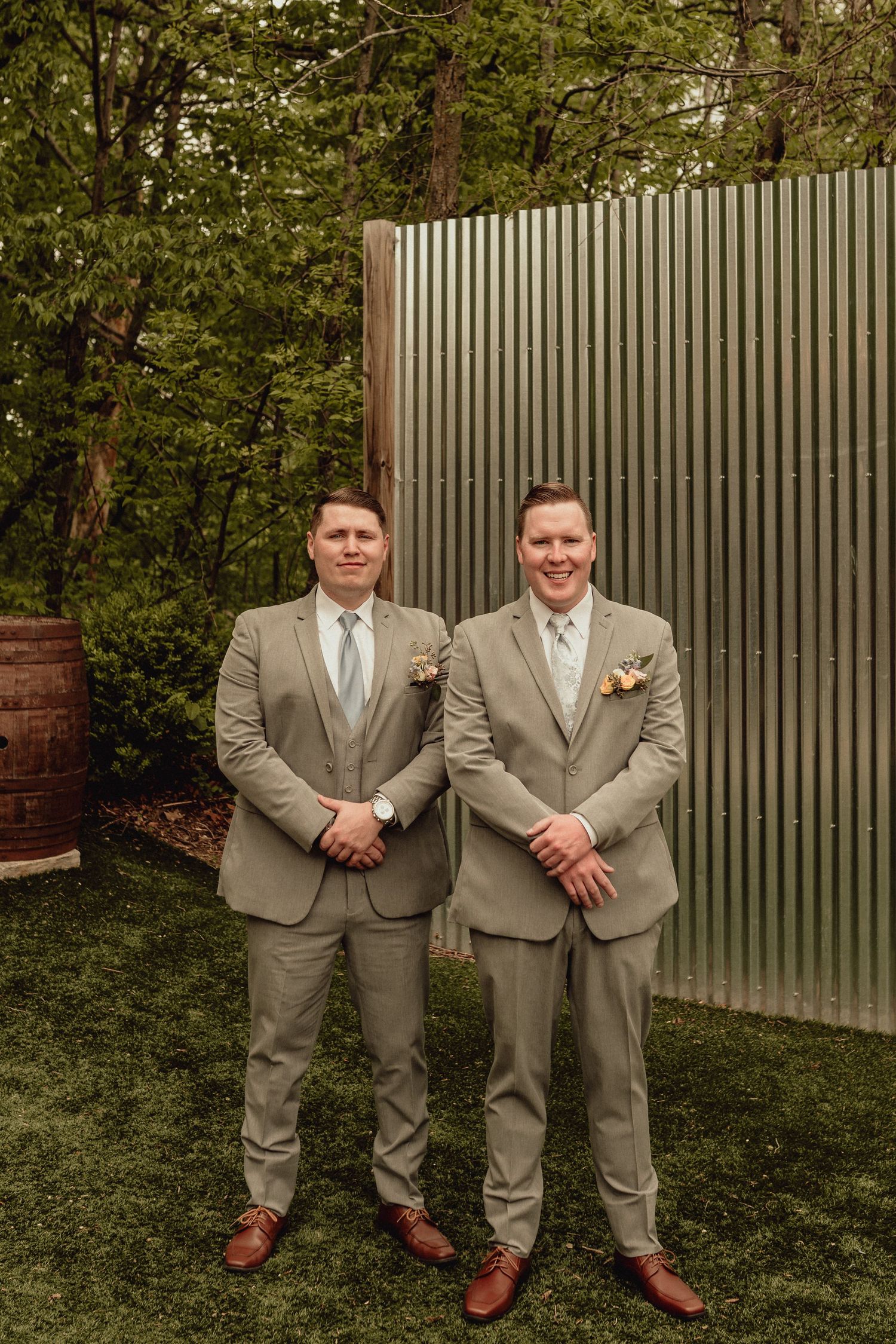 Two people in matching tan suits and ties stand together in a garden setting with a gray fence in the background.